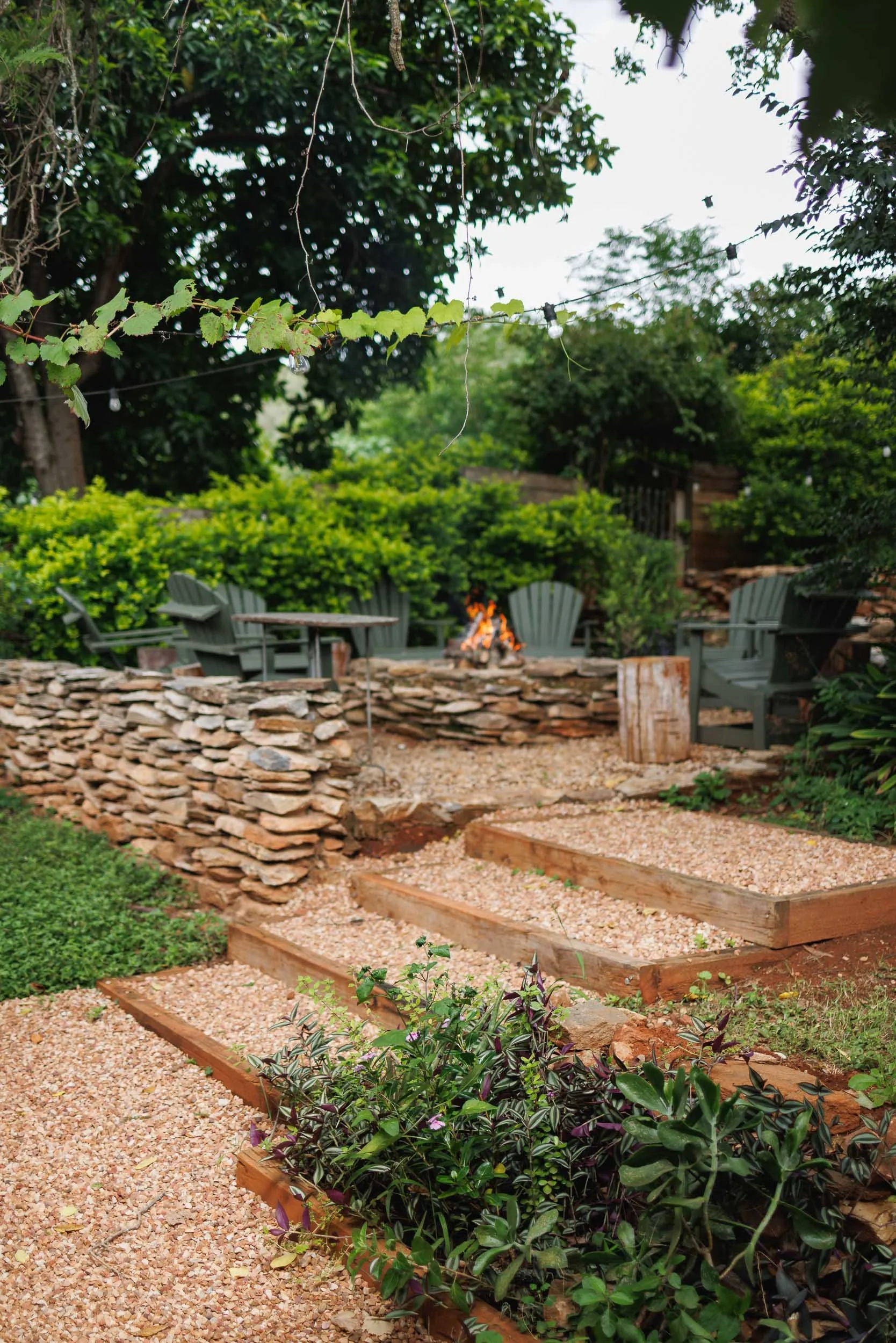 A backyard patio with a fire pit, surrounded by gray Adirondack chairs and lush greenery. String lights hang overhead, and there are raised garden beds with small plants in the foreground. A stone wall borders the patio area.