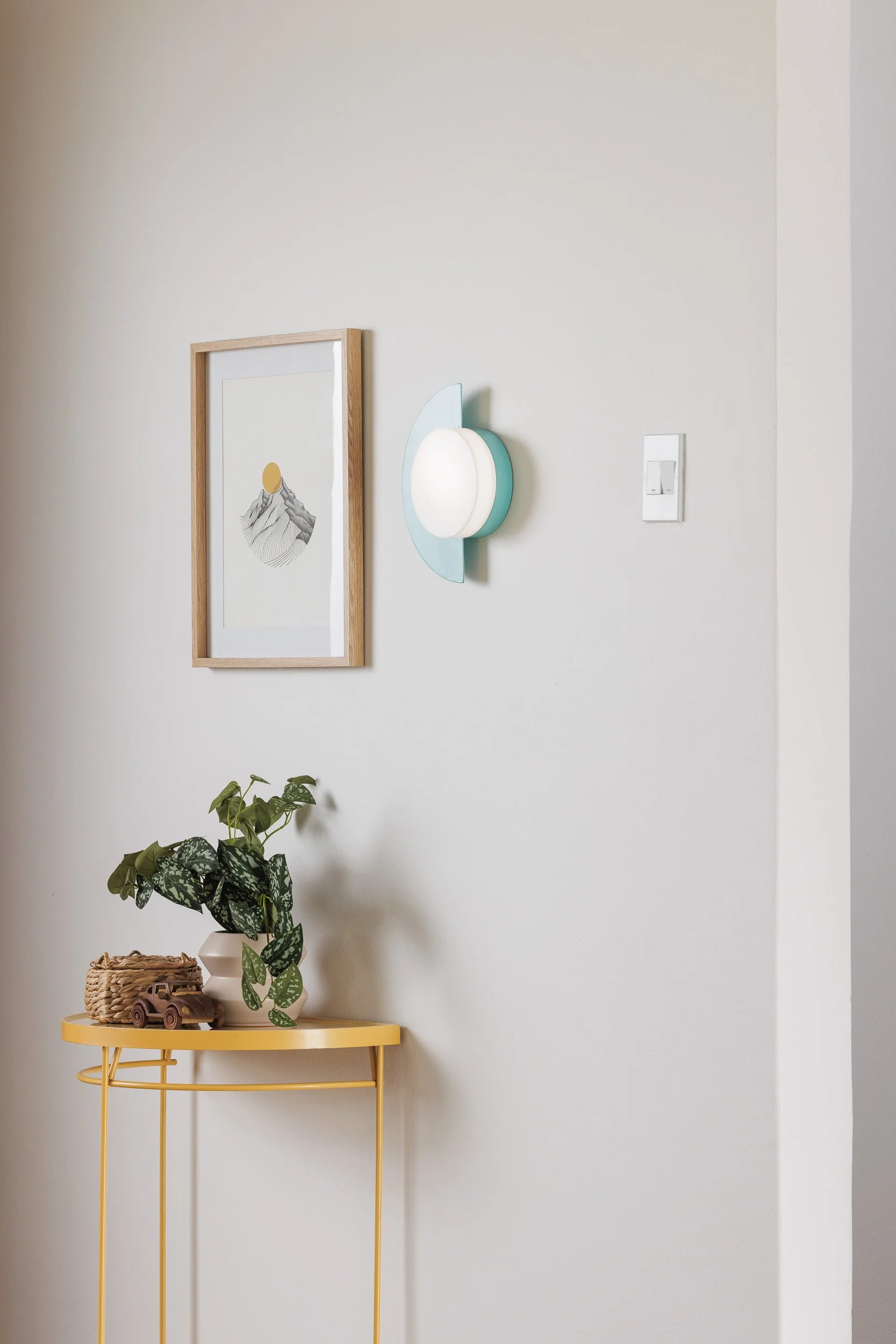Interior wall with framed art, wall light, and a yellow side table holding a potted plant, wicker basket, and toy truck.