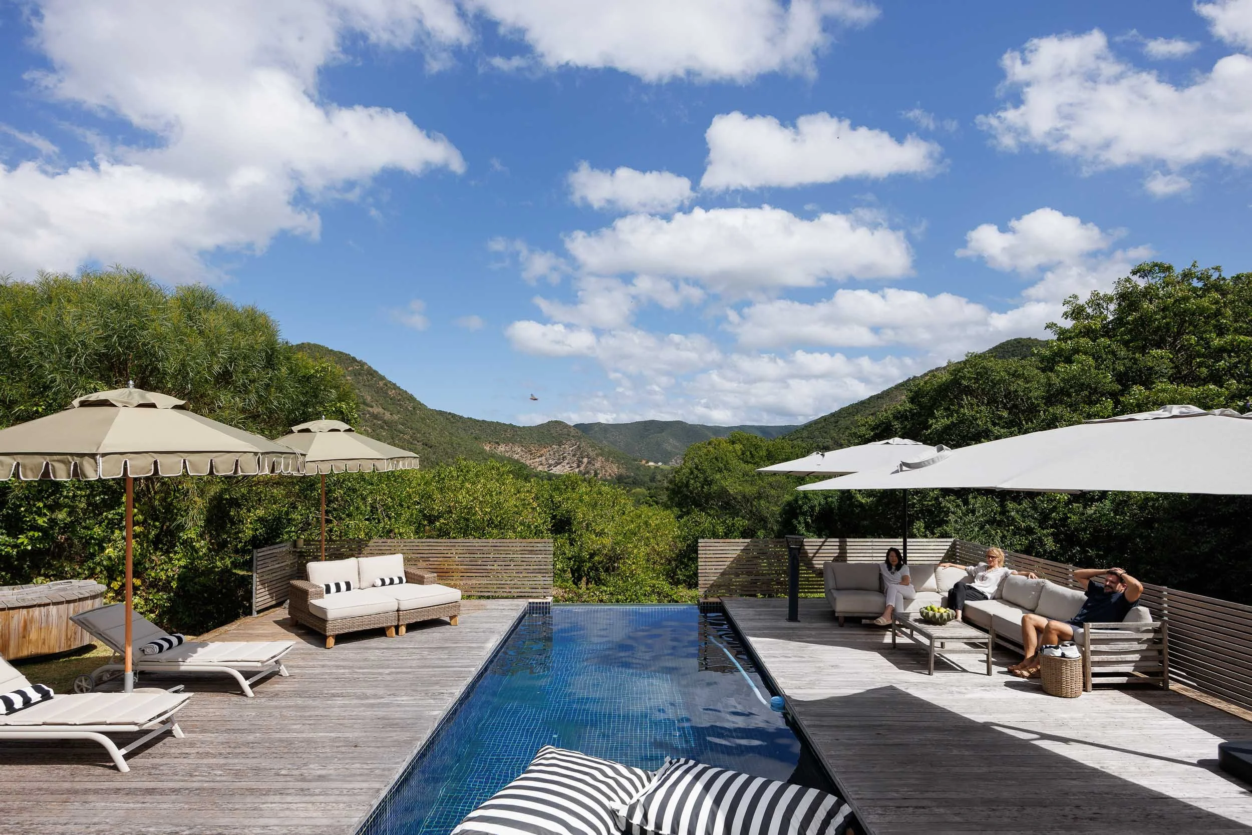 Outdoor pool area with lounge chairs, umbrellas, and people relaxing on couches, surrounded by green hills under a partly cloudy sky.