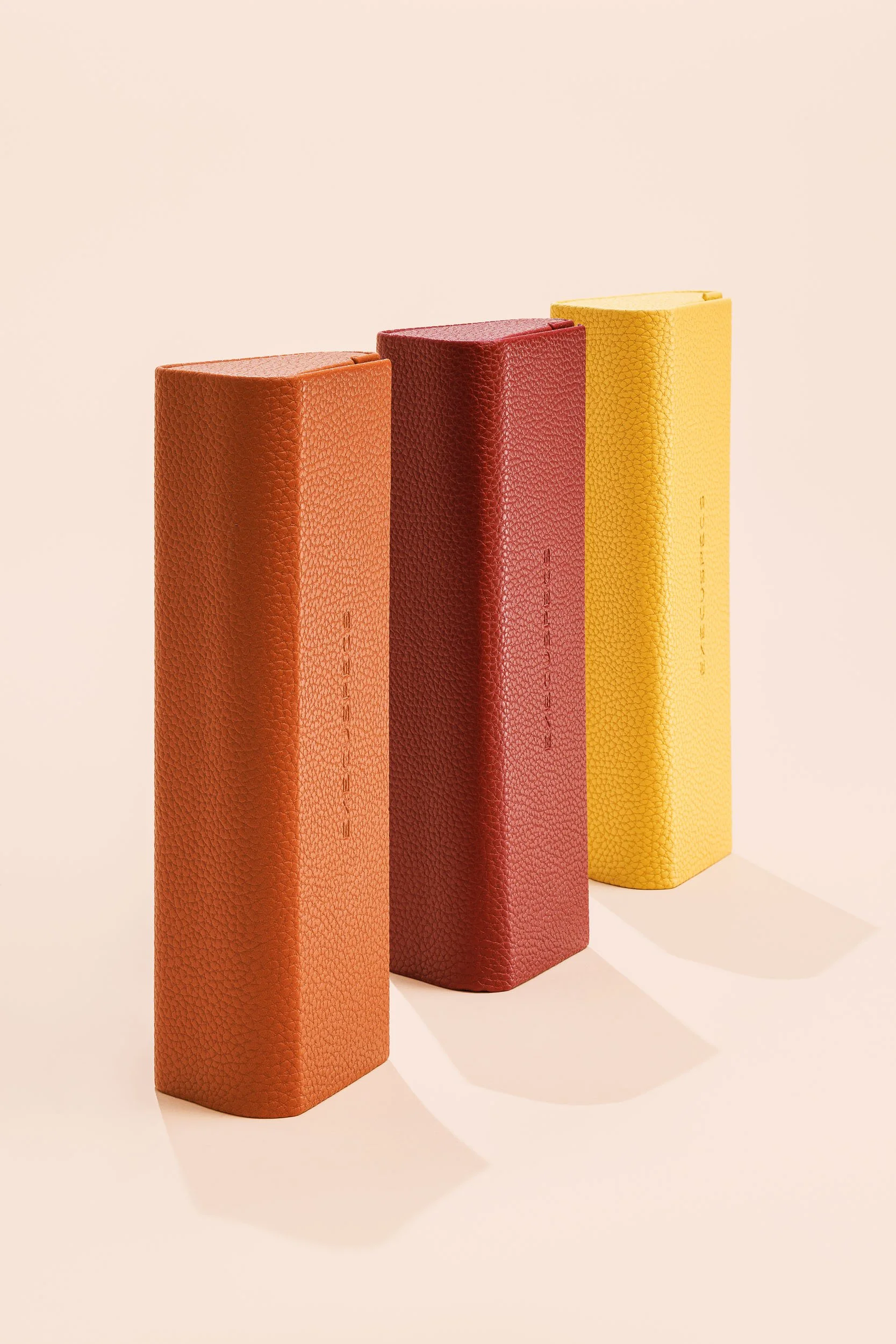 Three textured rectangular books in orange, red, and yellow, standing upright on a plain surface against a light background.