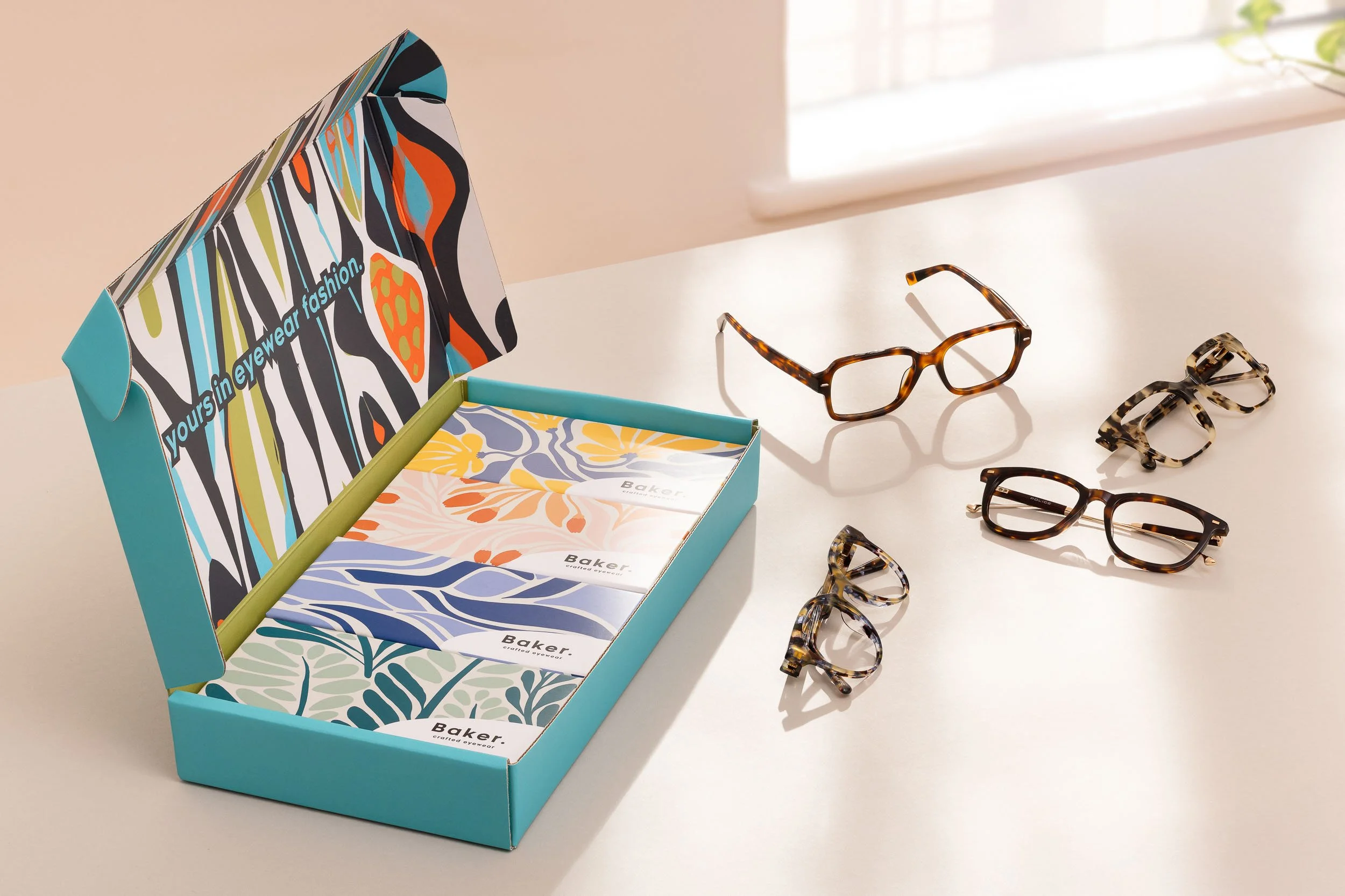 Open box of colorful, patterned eyeglass cases and a variety of tortoiseshell eyeglasses on a white table near a window.