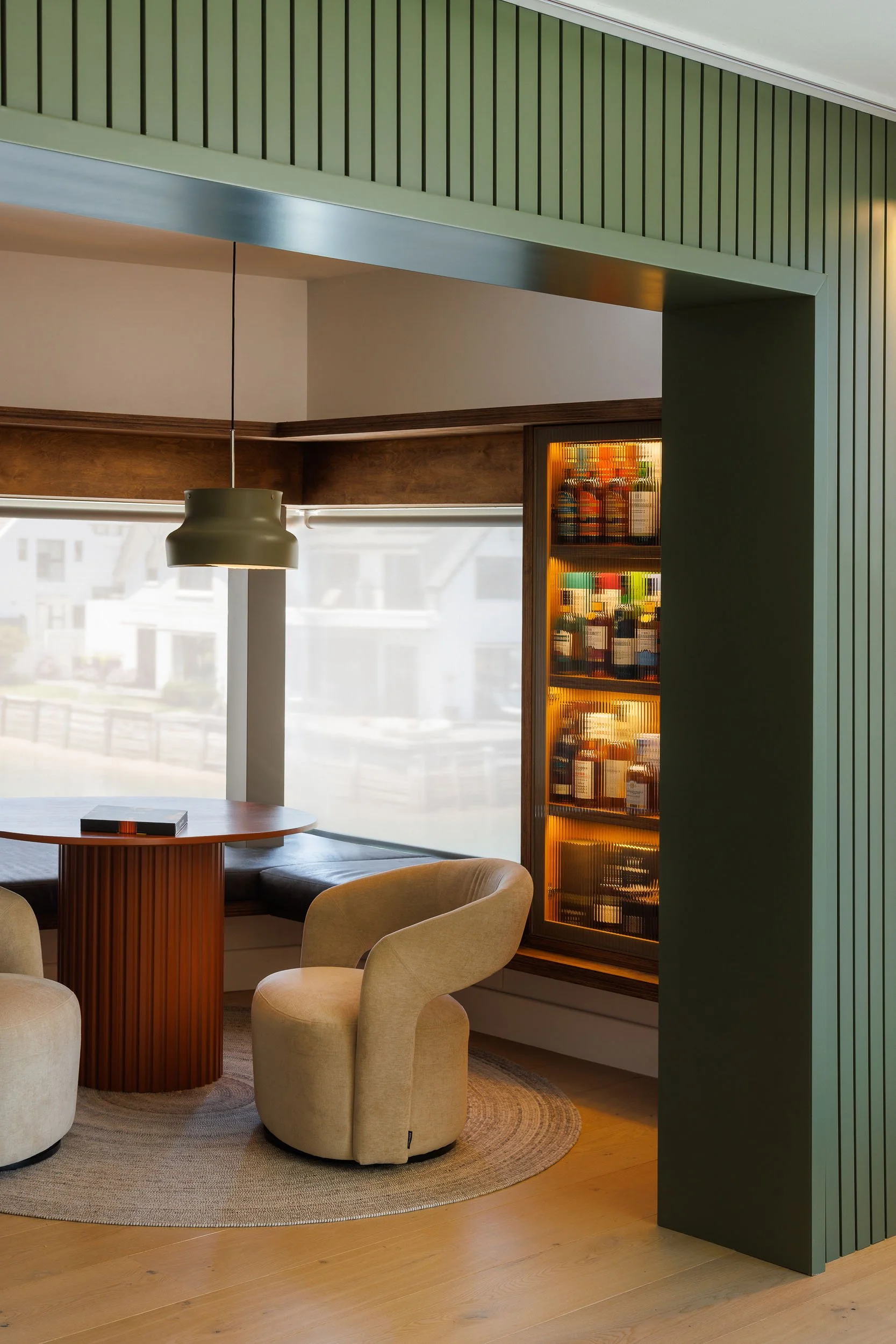 Intimate lounge nook with sage green slatted walls, backlit whisky display, and curved cream armchairs