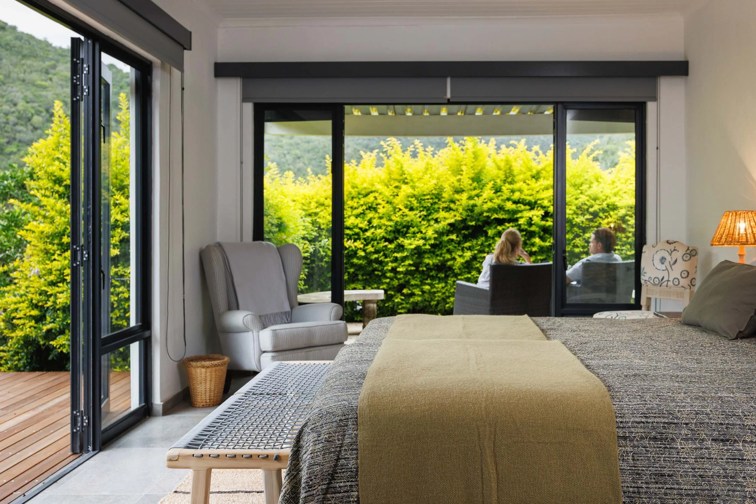 Living room with large glass sliding doors opening to a deck with greenery outside. Inside, there are chairs, a lamp, and a bed with blankets.