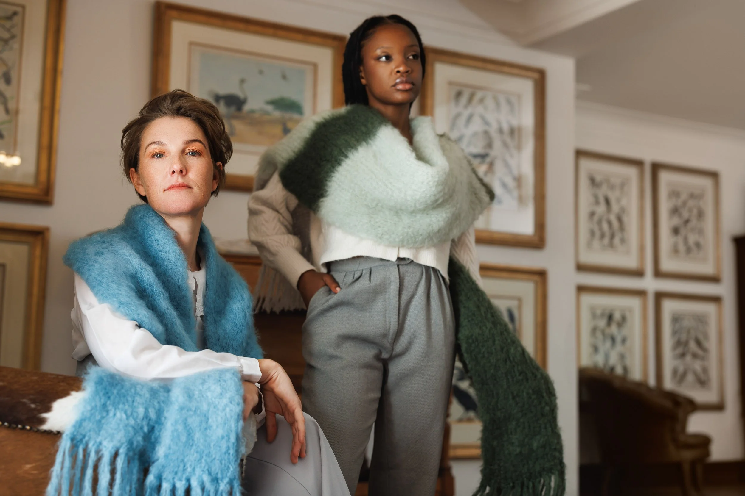 Two women in an art gallery, one sitting and the other standing, surrounded by framed artwork on the walls. The woman sitting has short hair and is wearing a blue fuzzy scarf, while the standing woman has long dreadlocks and is wearing a green fuzzy 