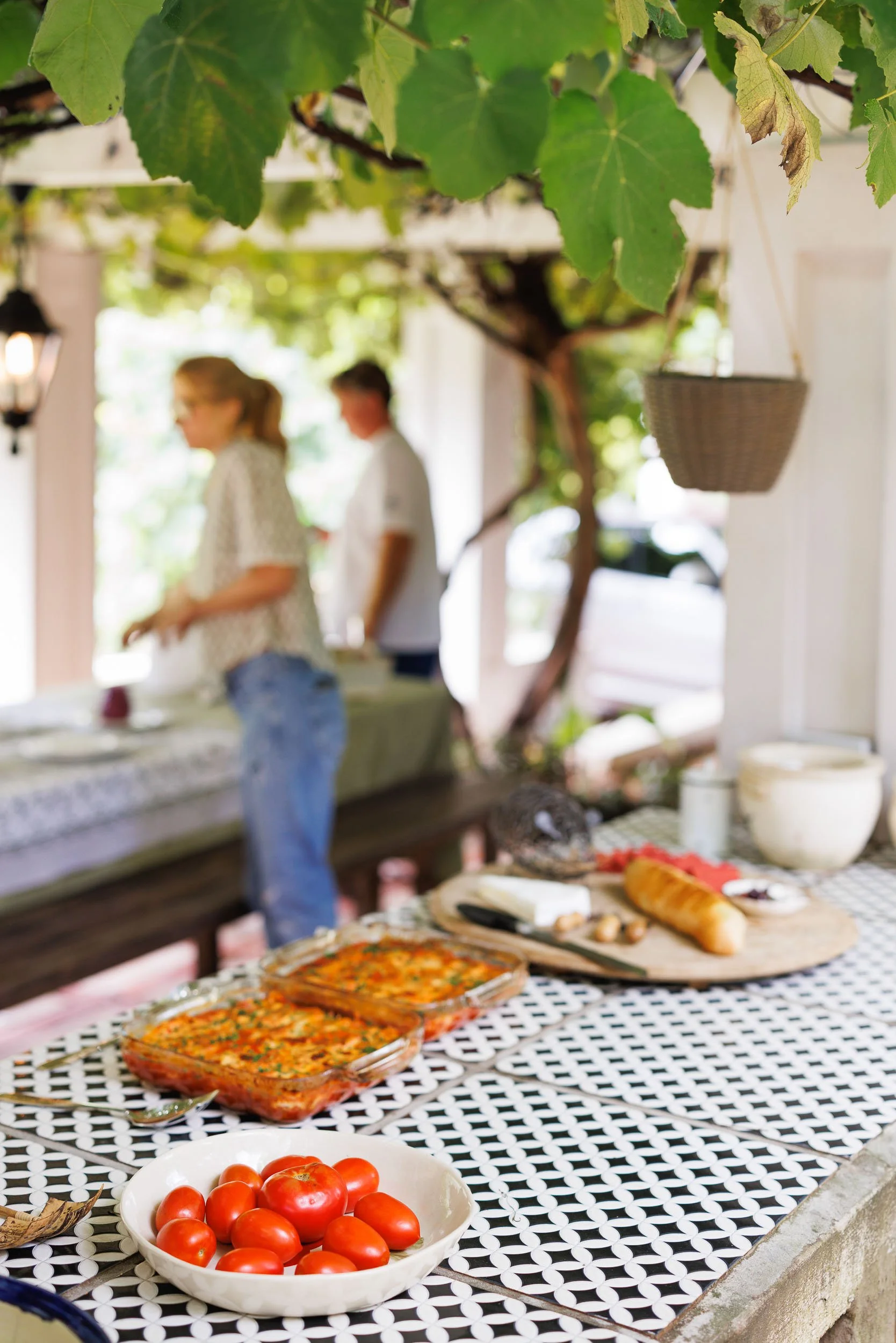 A table with various dishes of food, including a dish of tomatoes, baked dish, and bread, set outdoors under a tree with green leaves, with two people in the background at a buffet table.