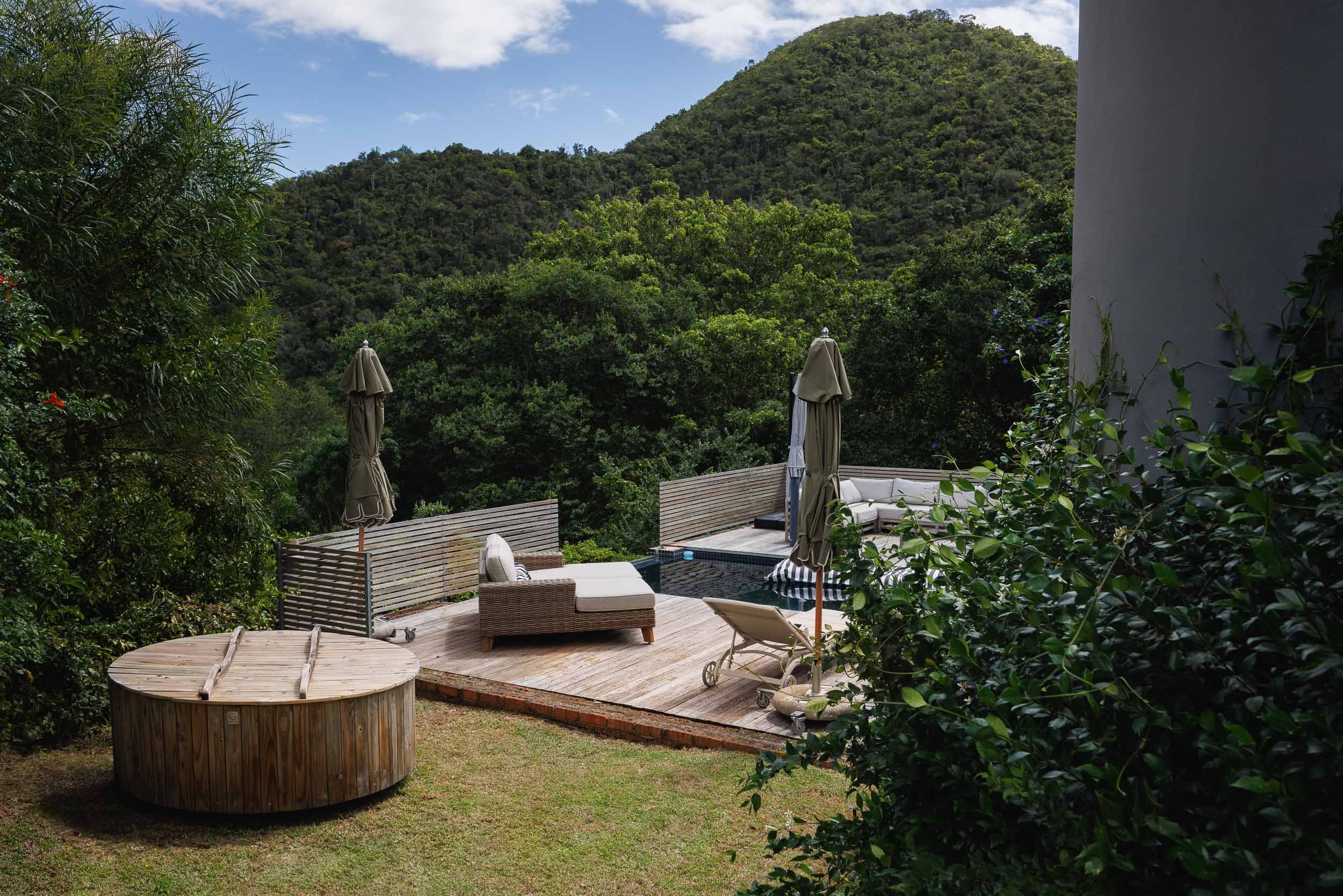 A backyard patio with outdoor furniture and umbrellas, surrounded by lush green vegetation and mountains in the background.
