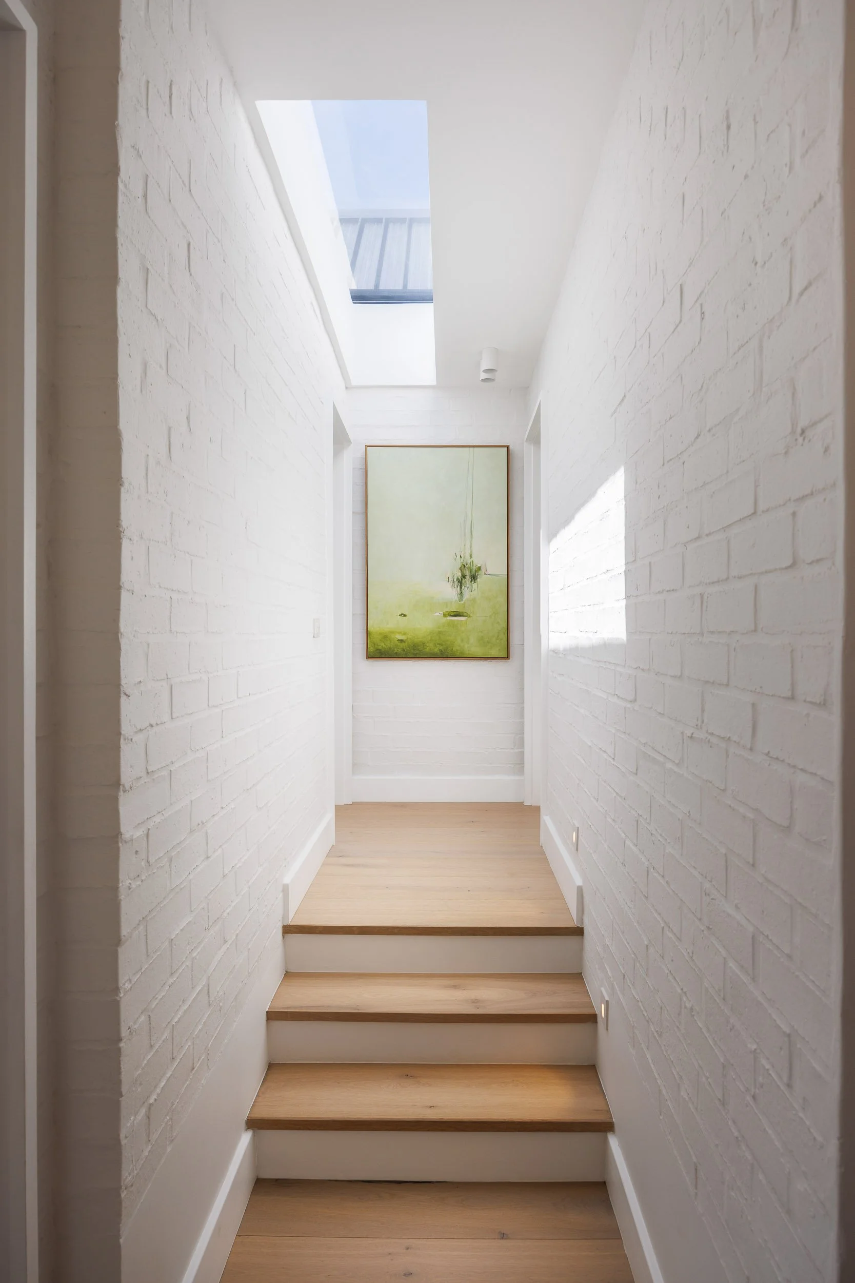 Narrow hallway with white-painted brick walls, skylight above, timber stair treads, and green landscape painting