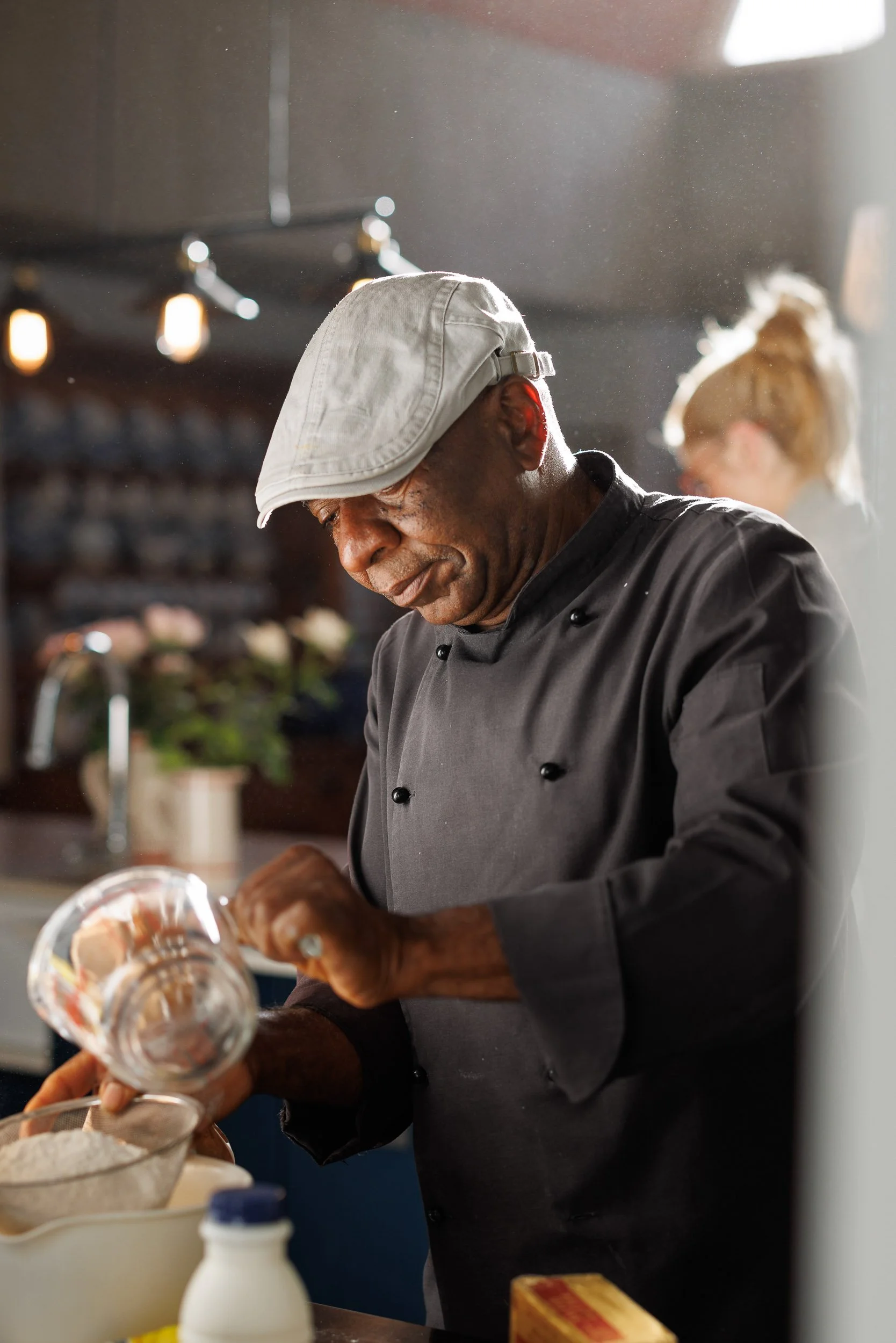 A chef wearing a beige hat and black chef uniform is preparing food in a kitchen, with a woman in the background and various kitchen items on the counter.