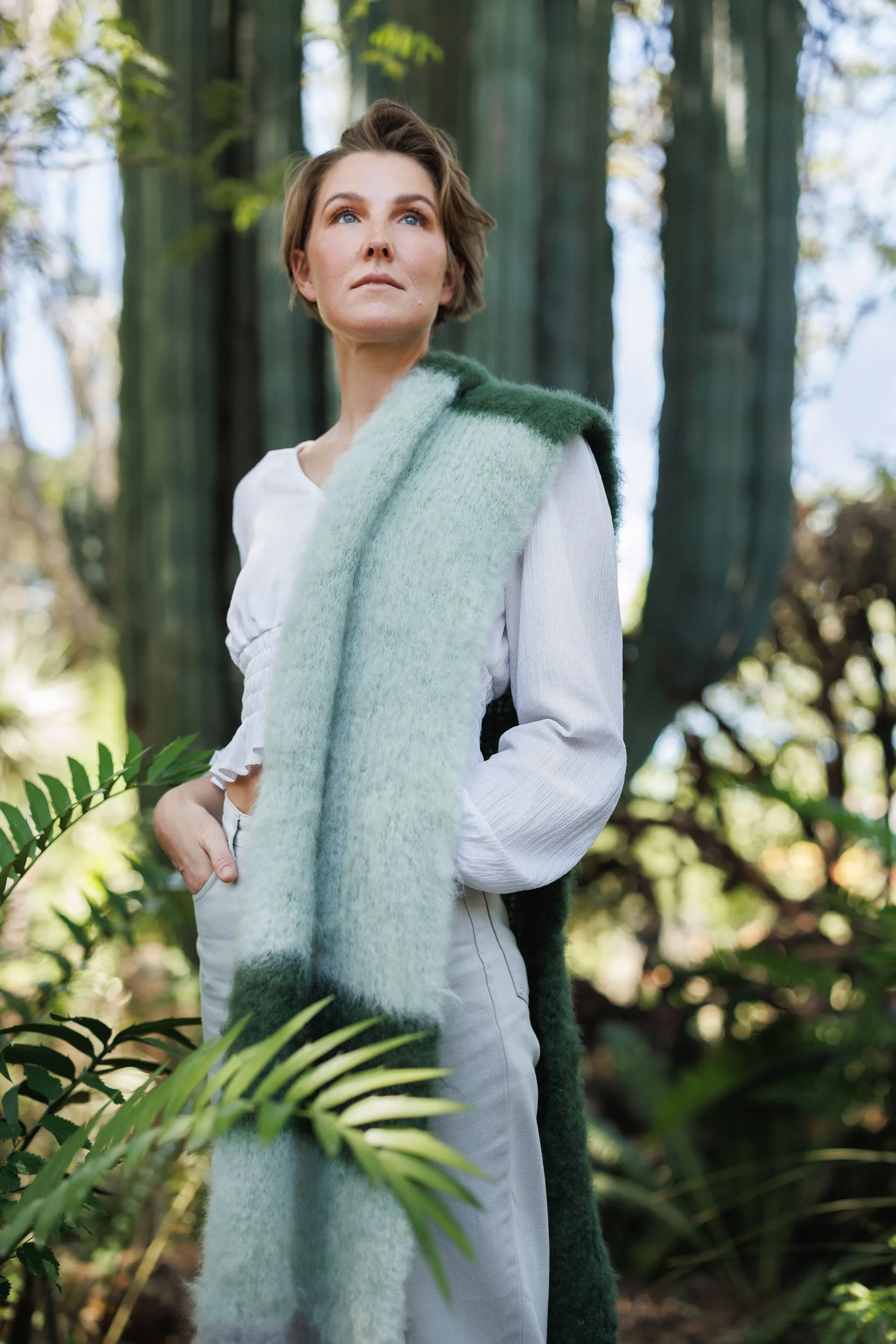A woman with short brown hair and blue eyes standing outdoors in a lush green forest, wearing a white long-sleeve shirt and a green and white fuzzy shawl draped over her shoulder, looking off into the distance.