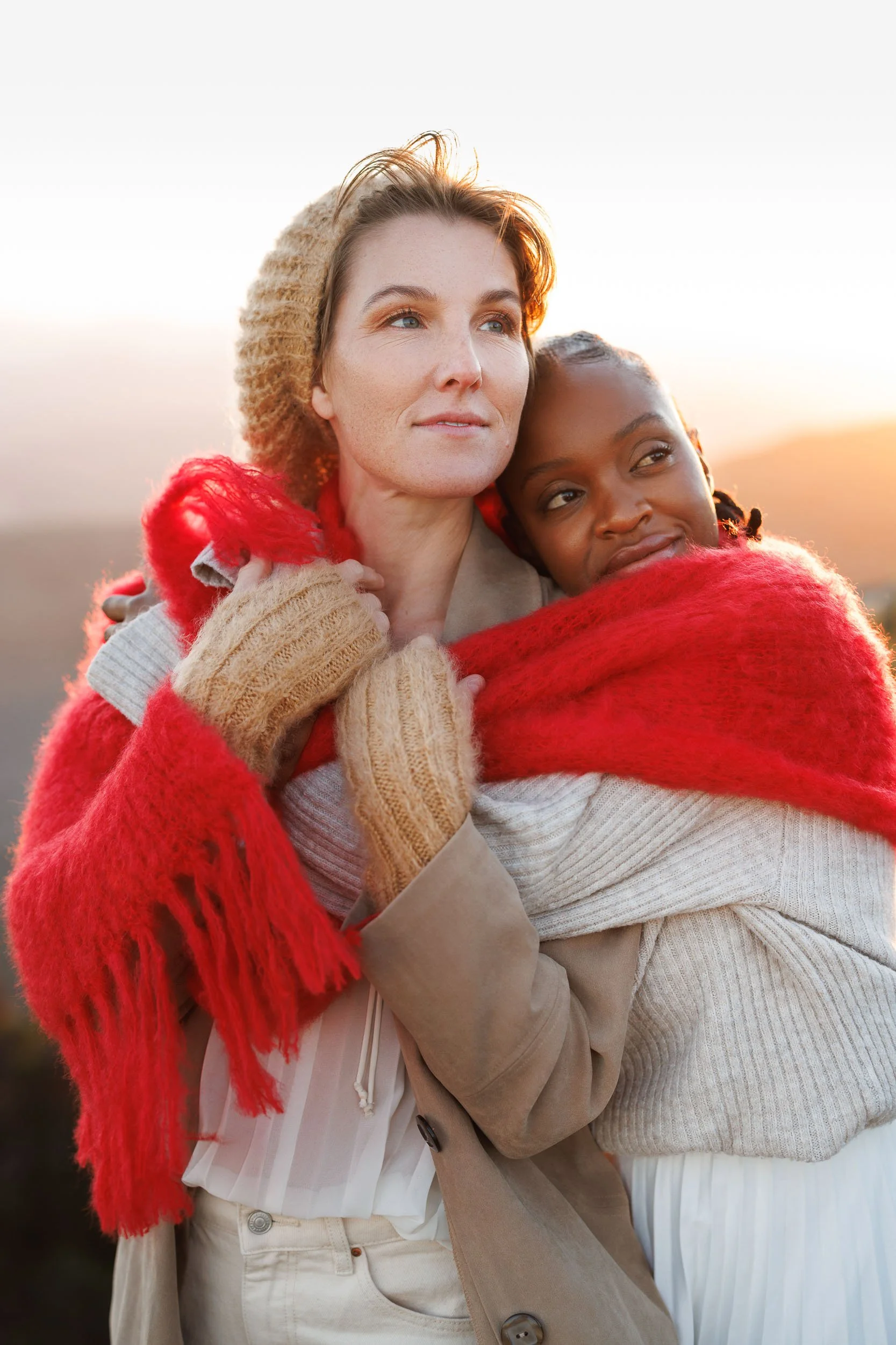 Two women embracing, one with blonde hair and the other with dark skin, outdoors at sunset.