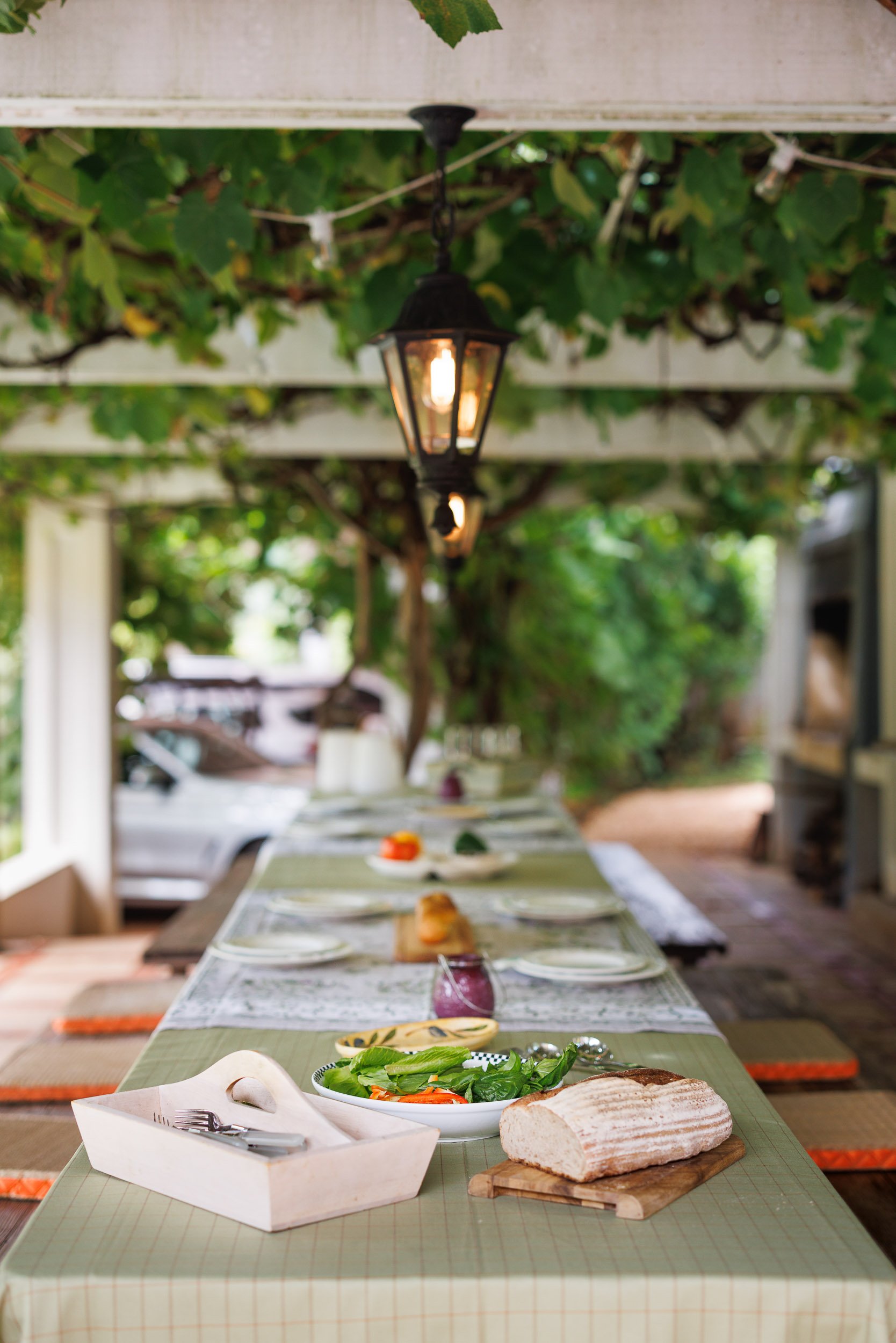 A long table set for a meal outdoors, with plates, bread, salad, and utensils on a green tablecloth. Overhead, a black lantern hangs from a beam amidst greenery.