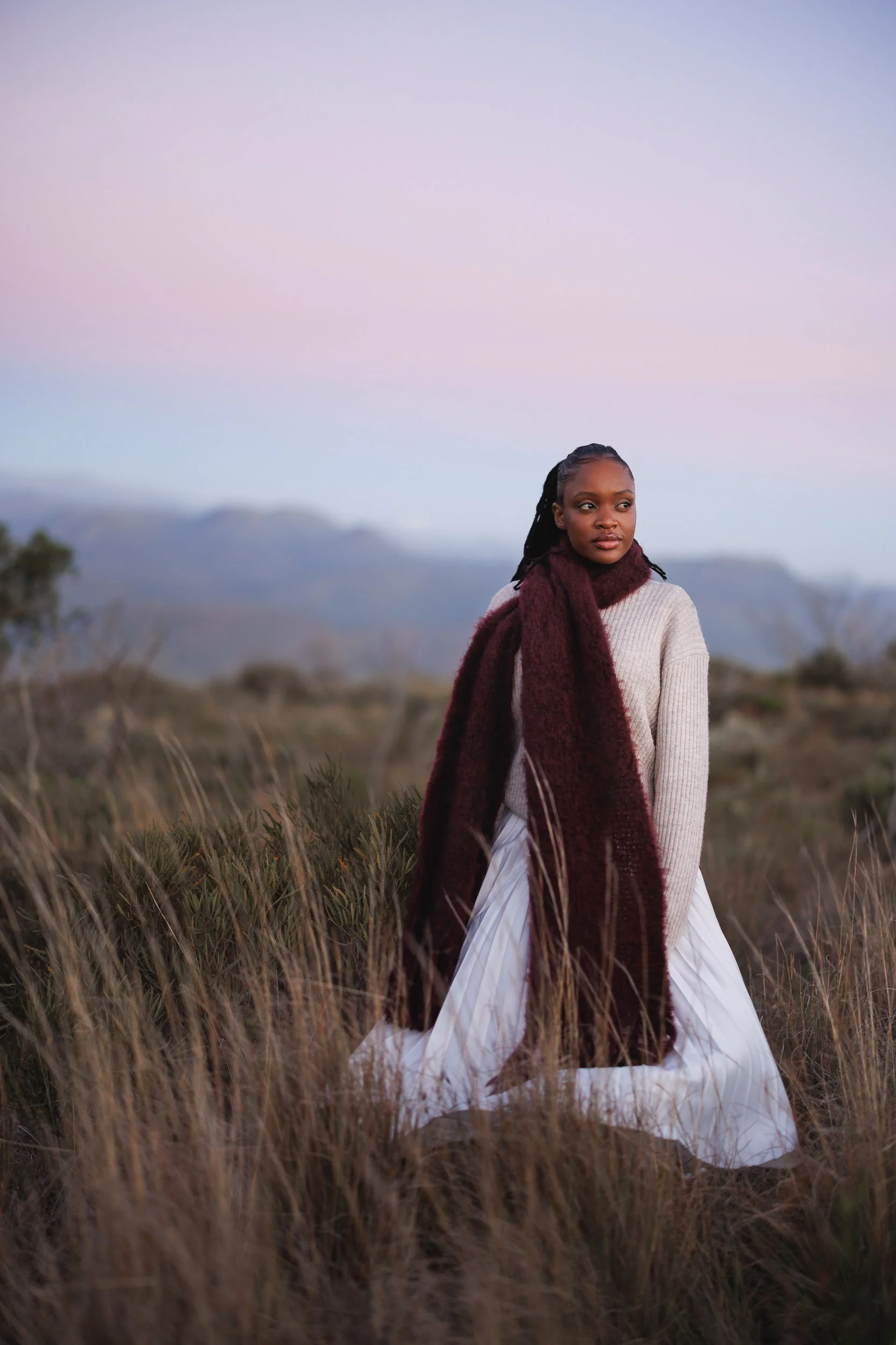 A woman with braided hair and dark skin standing in a field of tall grass during dusk, with mountains in the background and a pastel-colored sky.