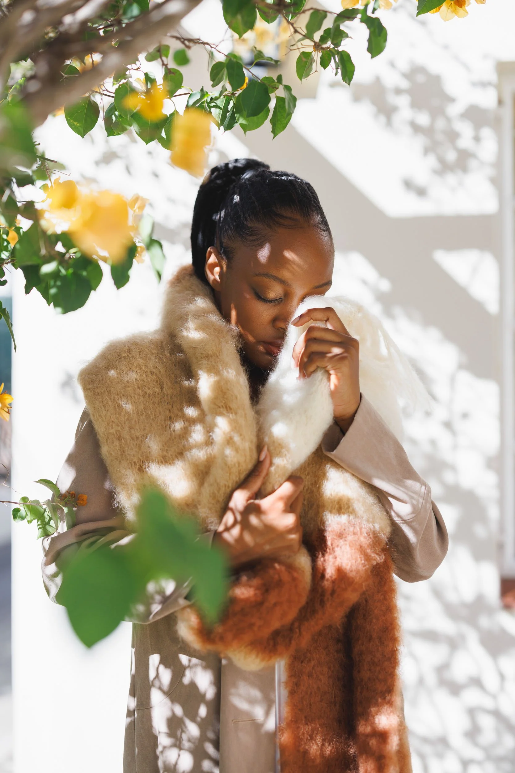A woman with braided hair wearing a fuzzy brown and cream coat, holding a soft white fluffy scarf, standing near a plant with yellow flowers and green leaves, with sunlight casting shadows on white walls.