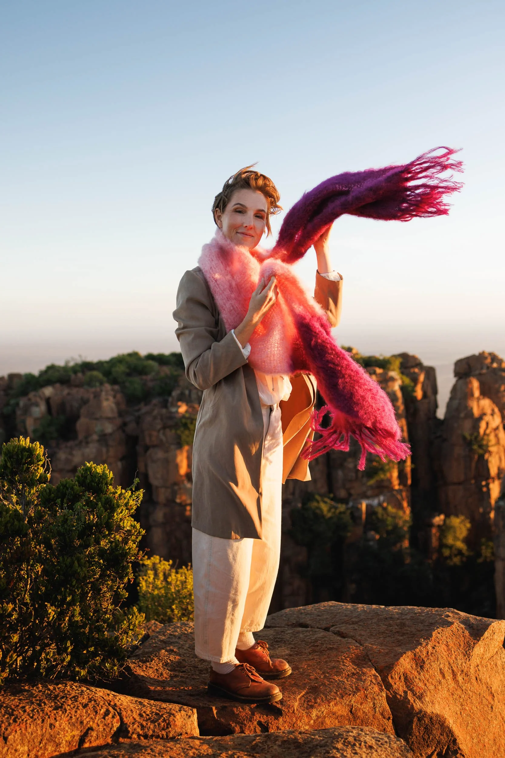 A woman with short hair wearing a beige jacket, light-colored pants, and brown shoes holds a large, colorful, fuzzy scarf that is pink, purple, and red. She is standing on rocky terrain and smiling at the camera during sunset, with cliffs and greener