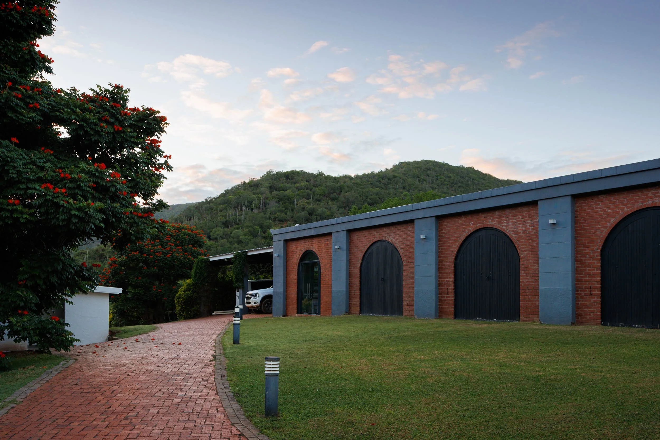 A brick building with arched black doors, a gray roof, and outdoor lighting fixtures. A brick pathway leads to the building, surrounded by a grassy lawn with short posts. To the left, a large tree with red flowers is visible. In the background, green