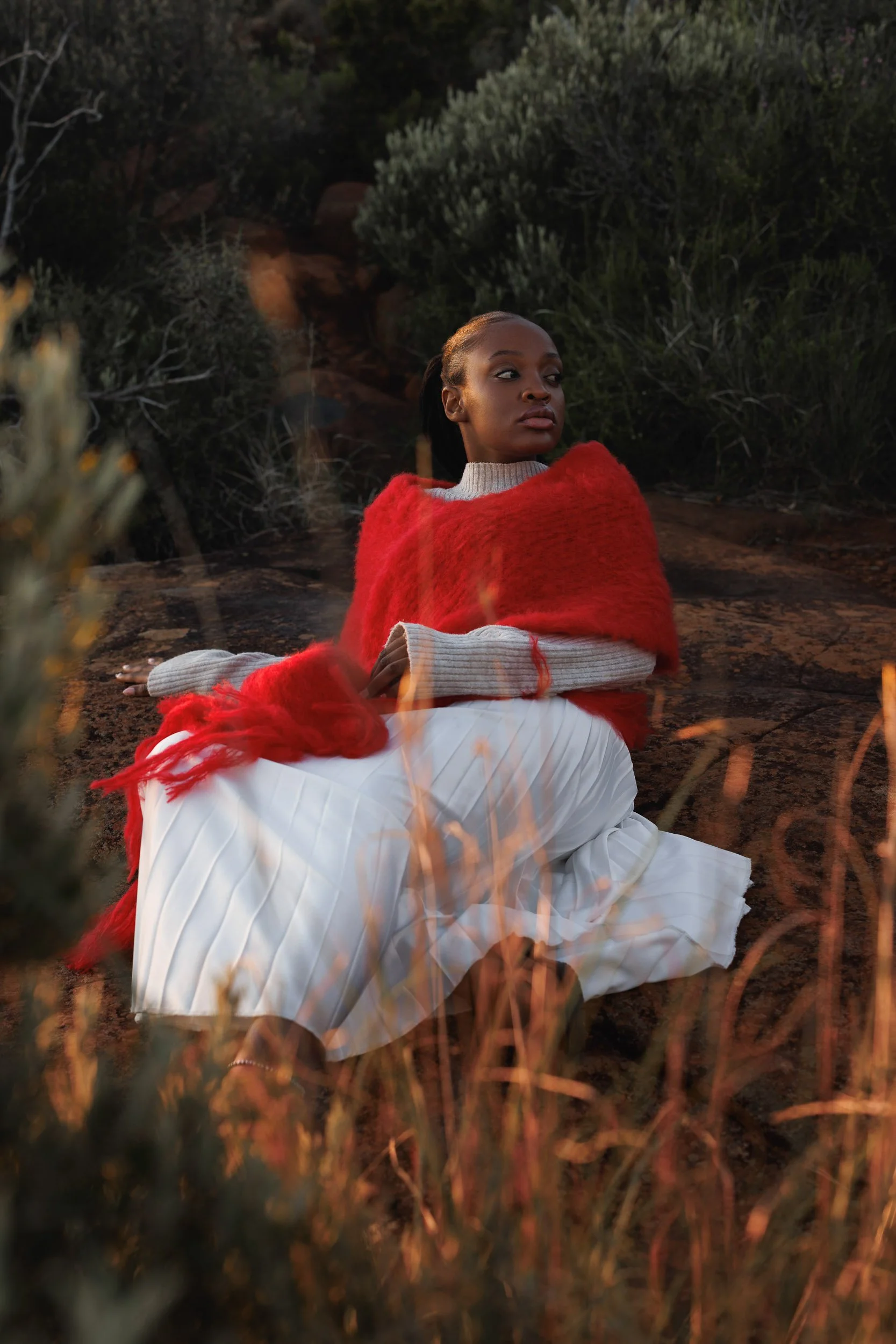A woman with dark skin and braided hair is seated outdoors on a rocky surface, wrapped in a red blanket, with a neutral expression, surrounded by bushes and natural vegetation during dusk.