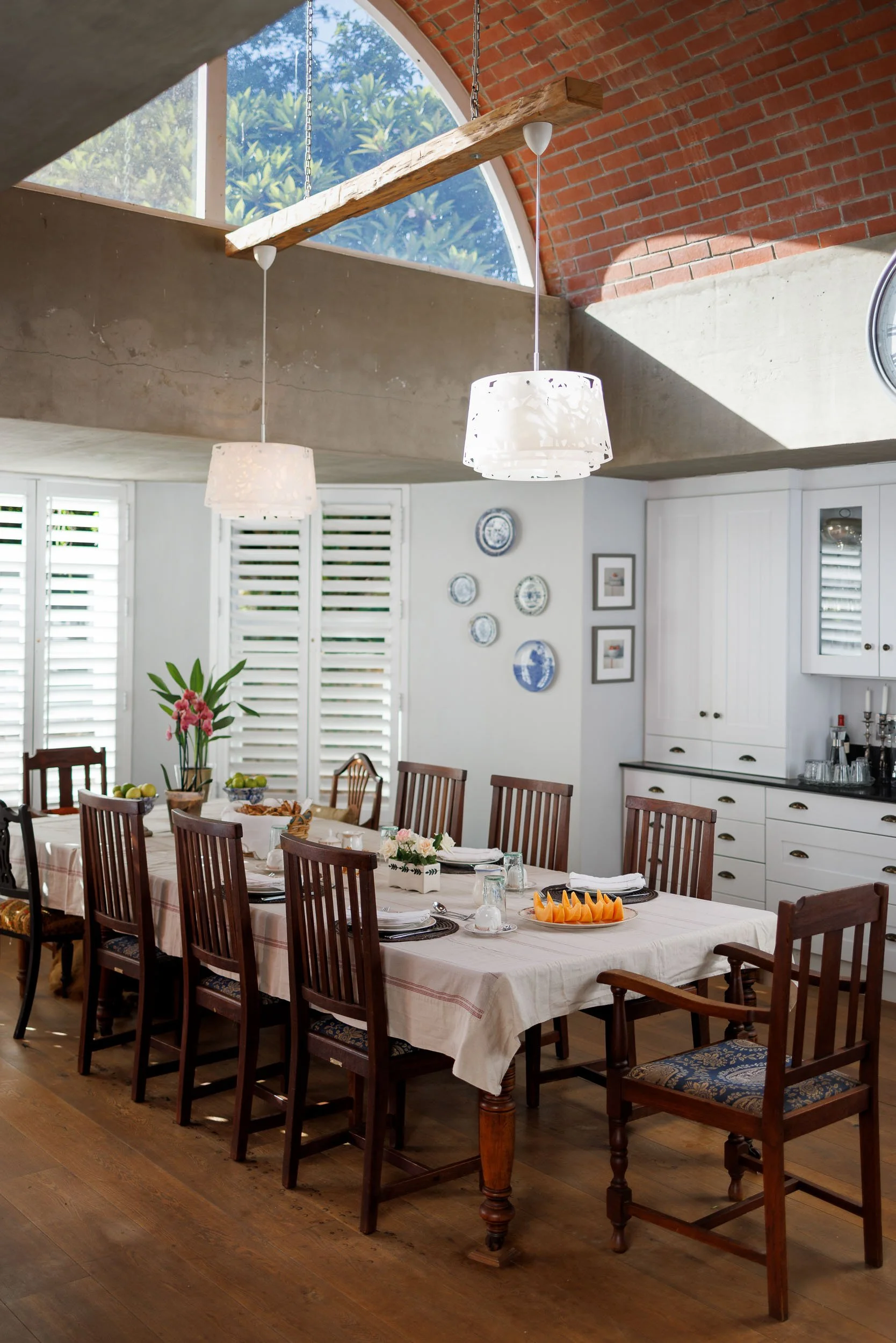 A dining room setup with a long table covered in a cream tablecloth, surrounded by wooden chairs with padded upholstered seats. The table is decorated with flowers and plates, and a platter of sliced fruit. The room features a brick and concrete ceil