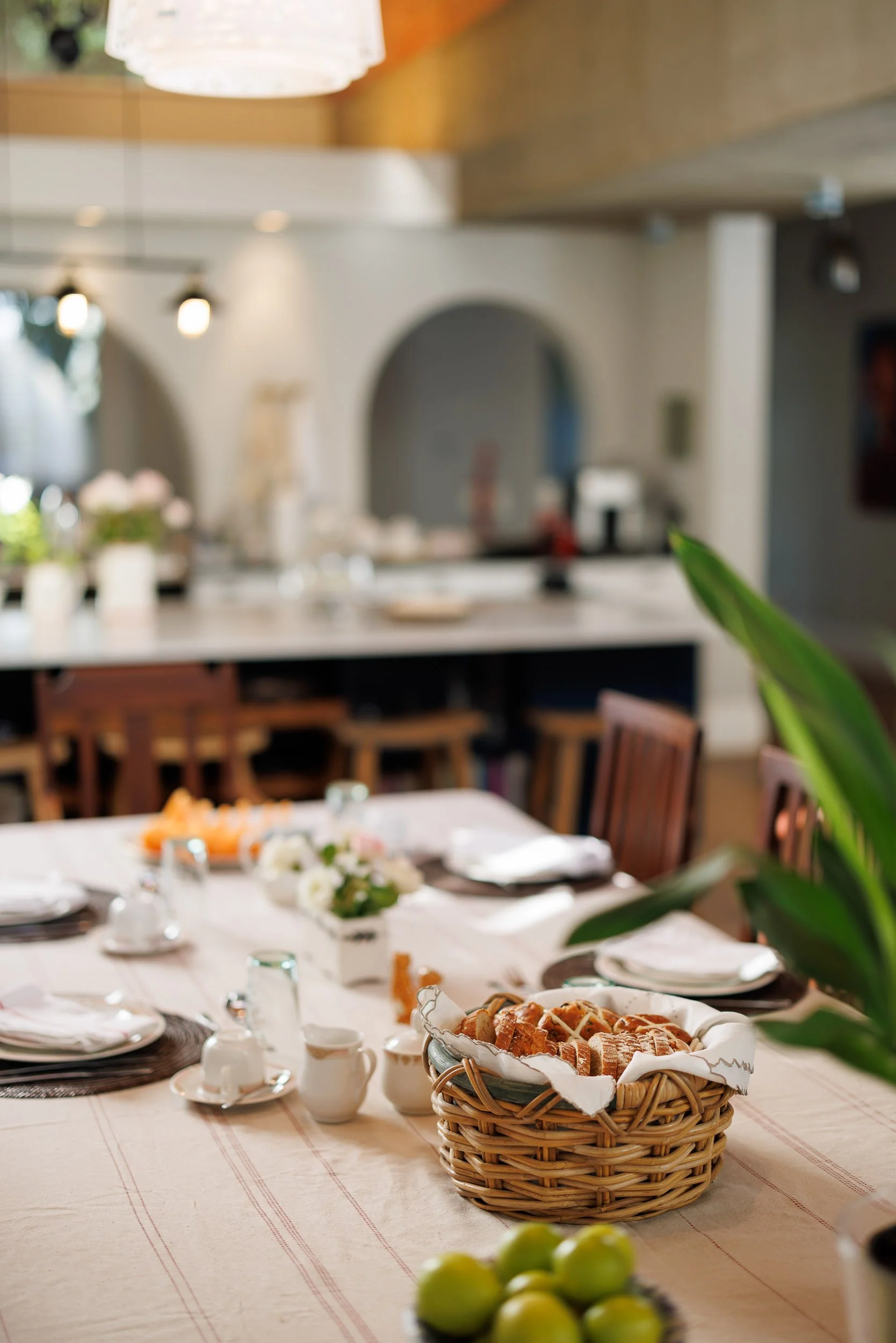 A dining table set for a meal with plates, cups, and a flower centerpiece, with a basket of bread in the foreground and a blurred background of a cozy dining room.
