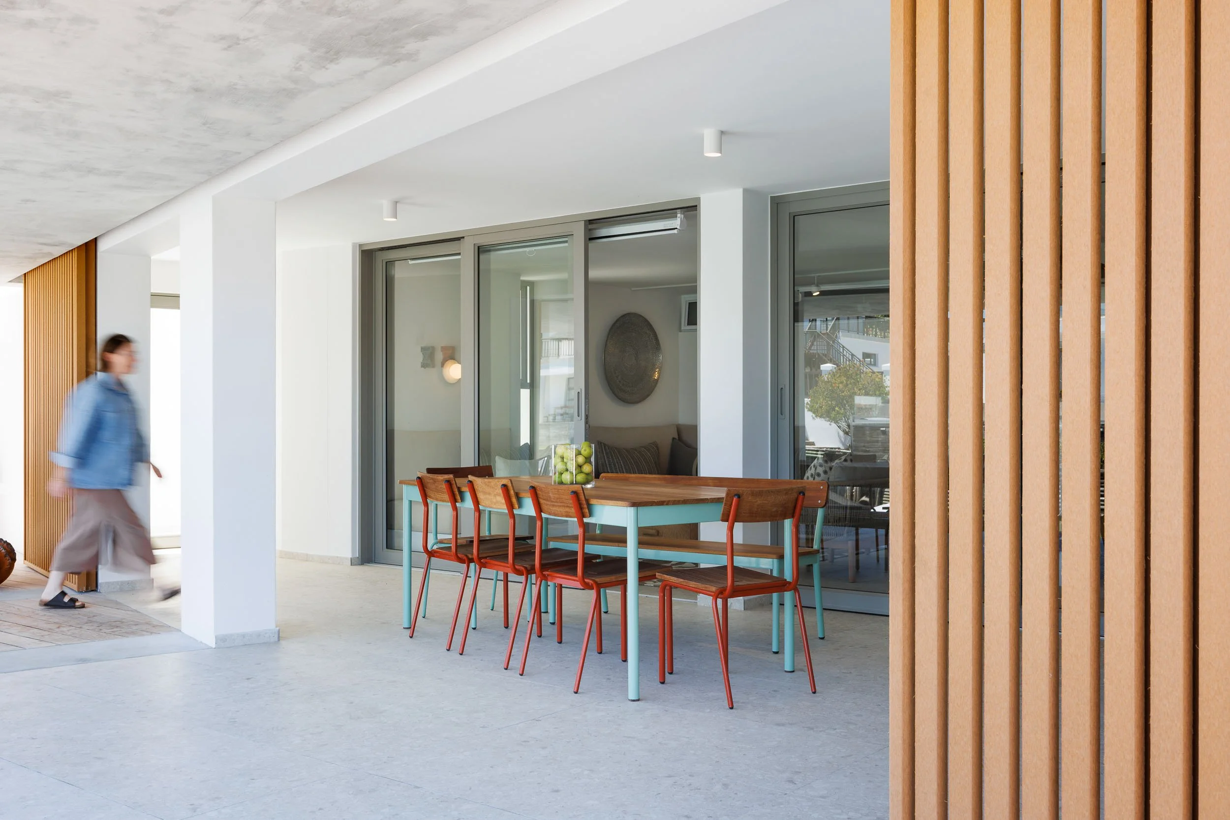 Shaded outdoor dining area with colourful red and teal metal chairs under concrete soffit, canal house beyond