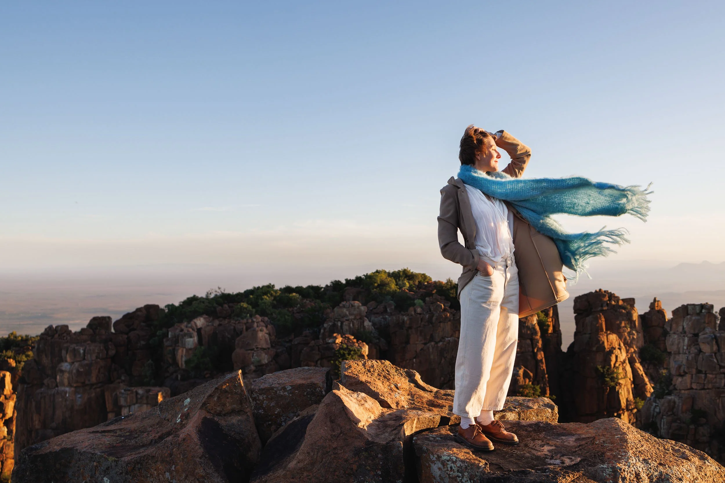 A woman standing on rocks at sunset, looking into the distance with her hand on her head, wearing a tan jacket, white shirt, light-colored pants, a large blue scarf, and brown shoes, with a landscape of rocky formations and a clear sky in the backgro