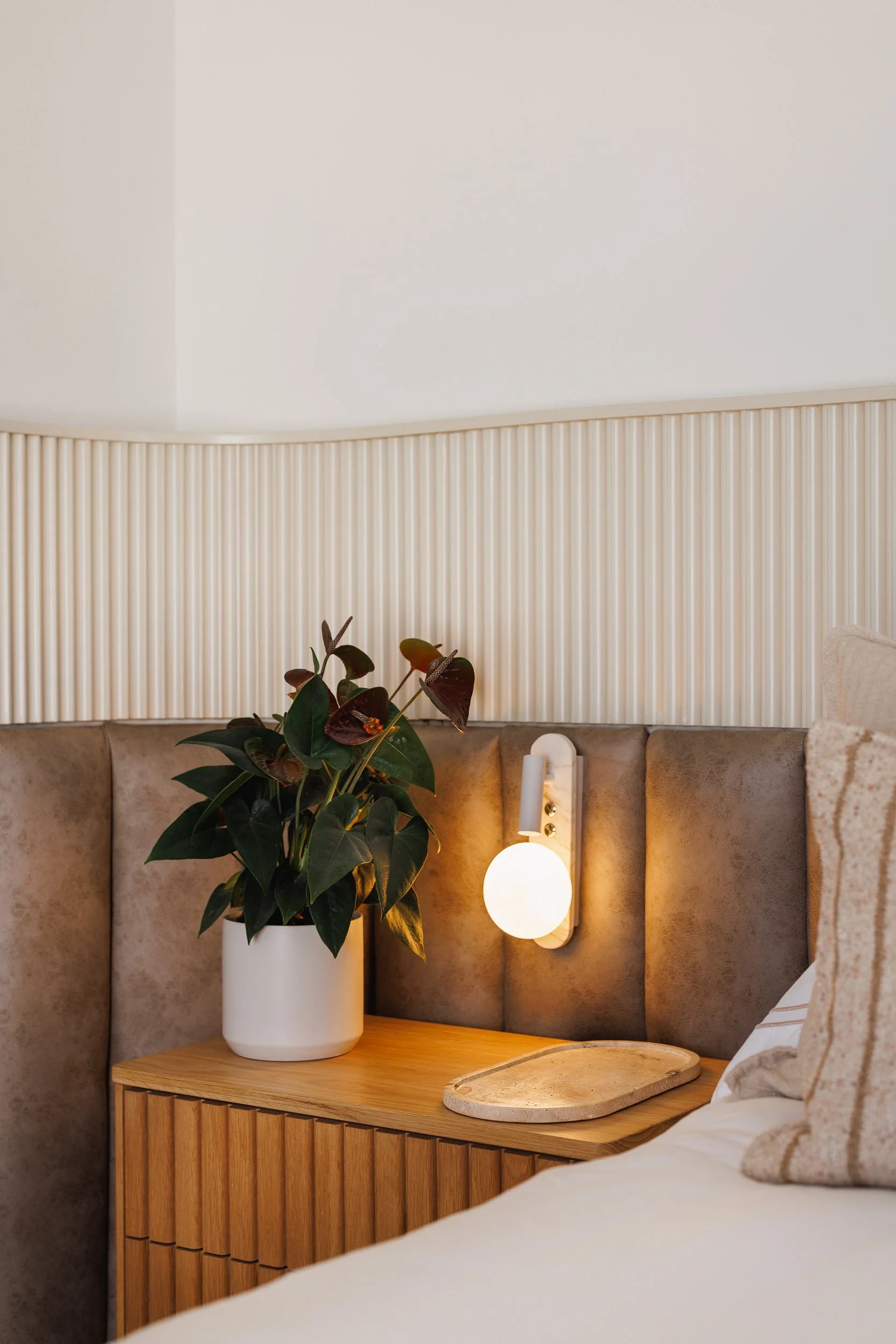 Bedside vignette with fluted timber nightstand, globe wall sconce, and dark-leafed anthurium plant