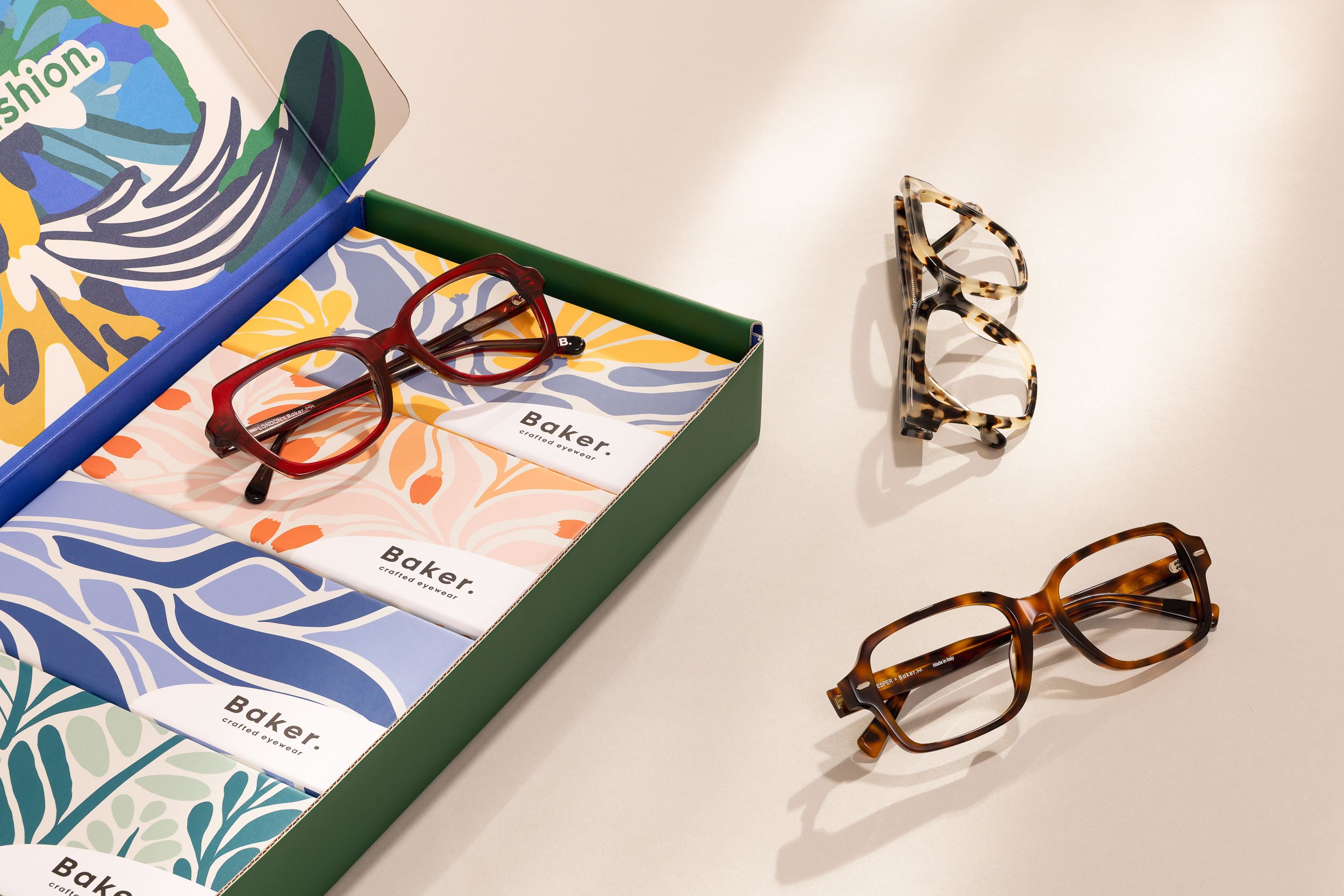 Several pairs of tortoiseshell and red eyeglasses on a white surface next to a colorful box labeled 'Baker Crafted Eyewear' with decorative patterns inside.