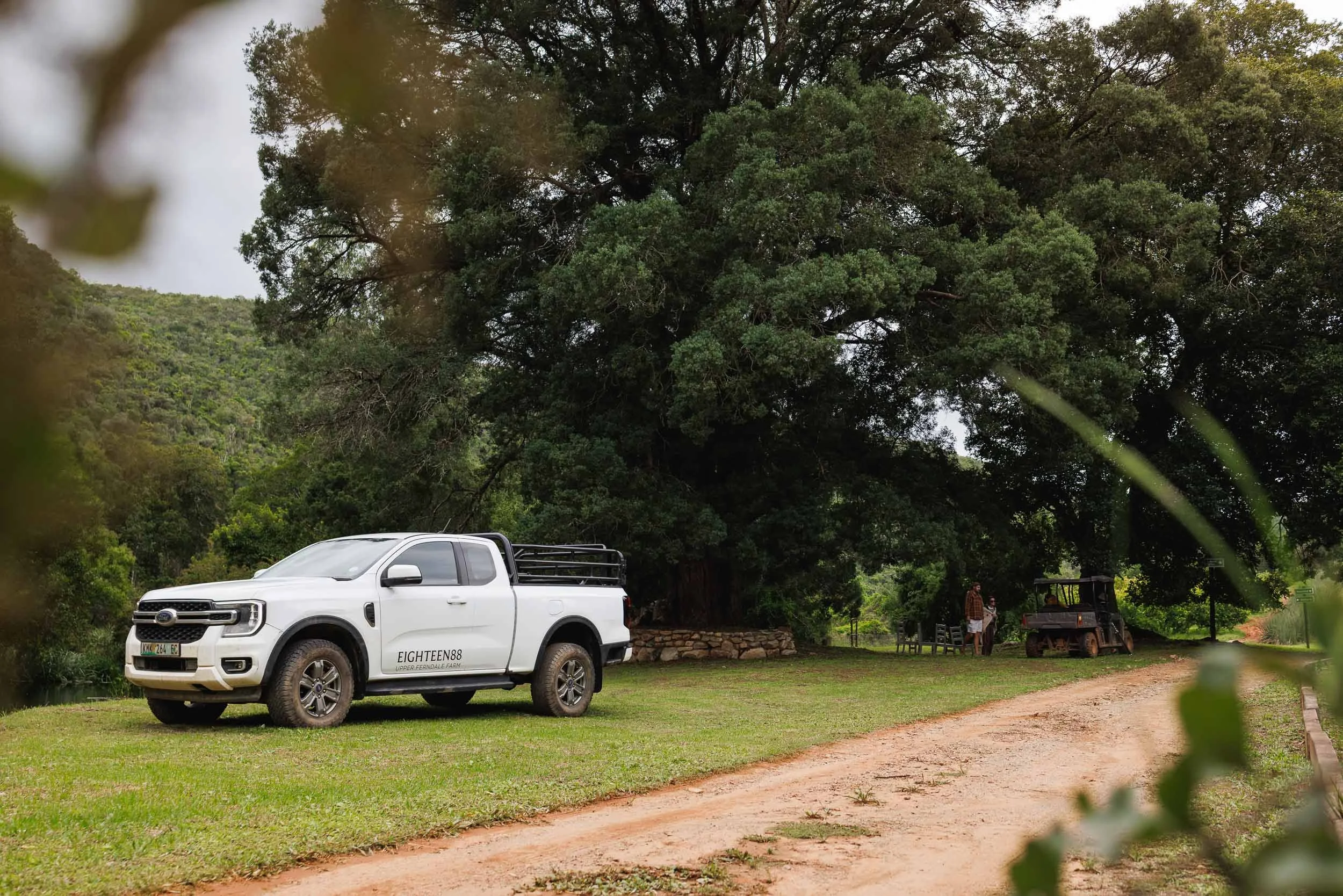 White pickup truck parked on grass near dirt path in a rural area with large trees and a hillside in the background. Two people and a utility vehicle are visible in the distance.