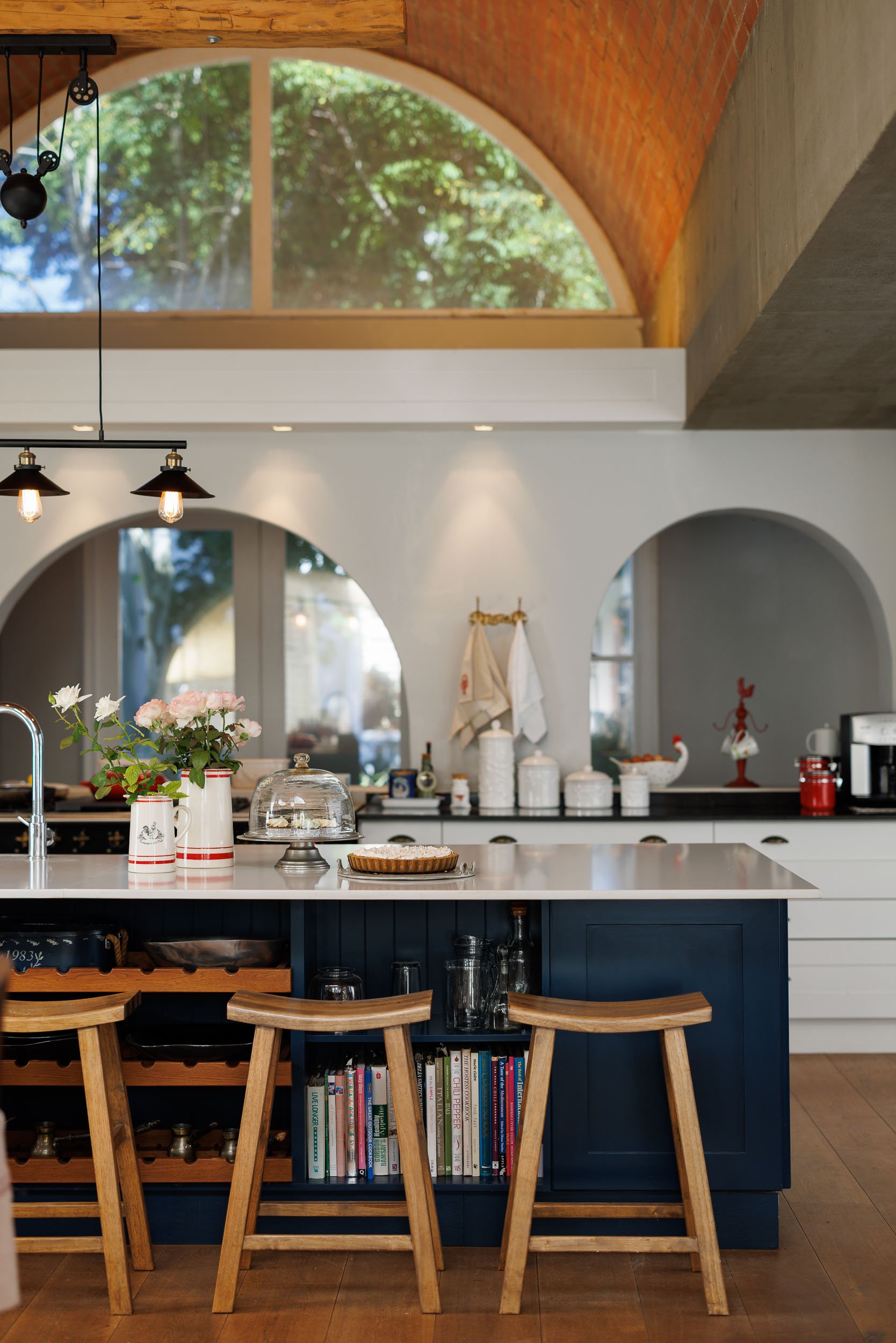 A modern kitchen with a white island counter, dark blue cabinetry, and wooden bar stools. The background has large arched windows, a high brick ceiling, and decorative items like flowers, books, and kitchen jars.