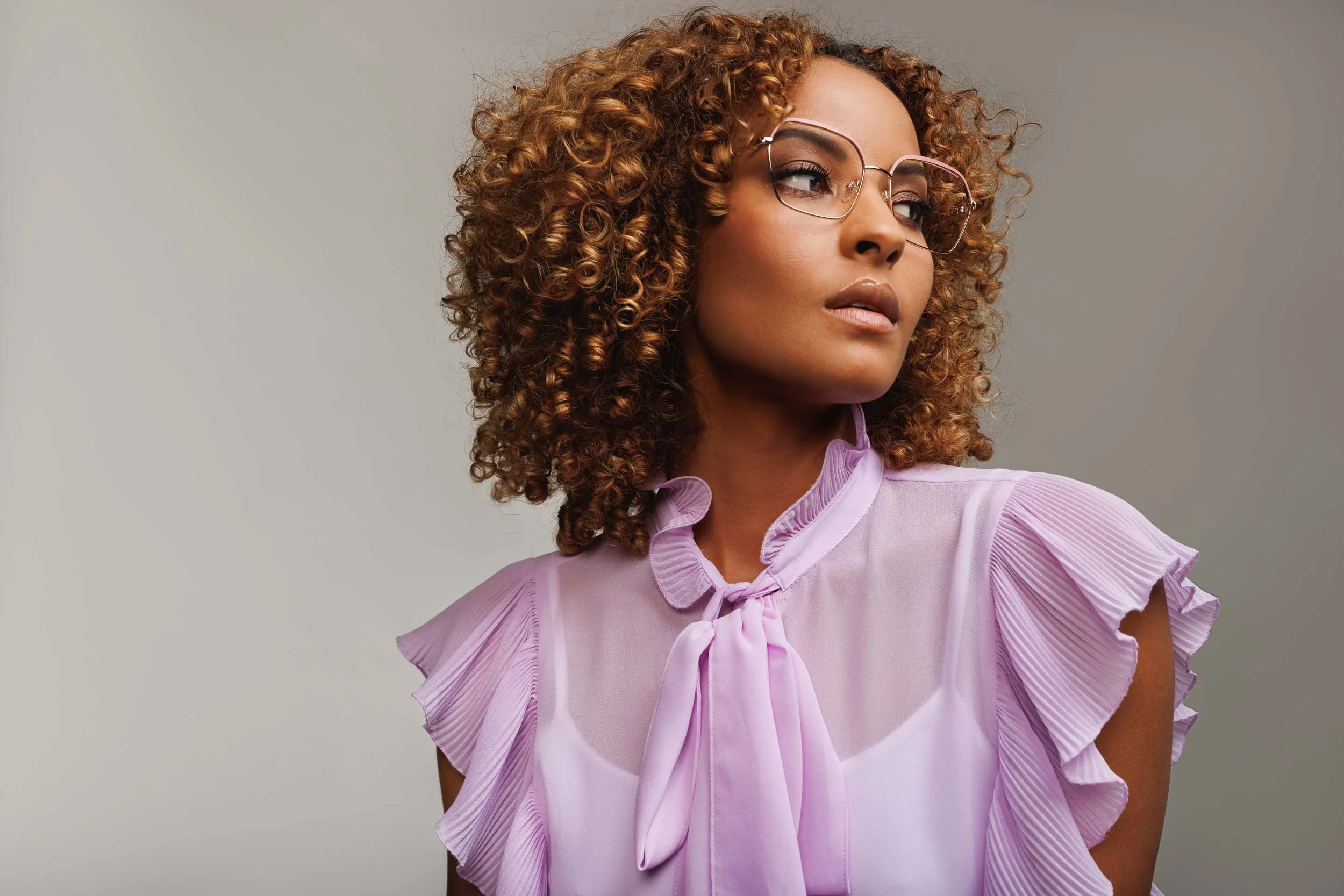 Studio portrait of young mixed race female wearing pink framed glasses and a sheer pink top with frilly sleeves.