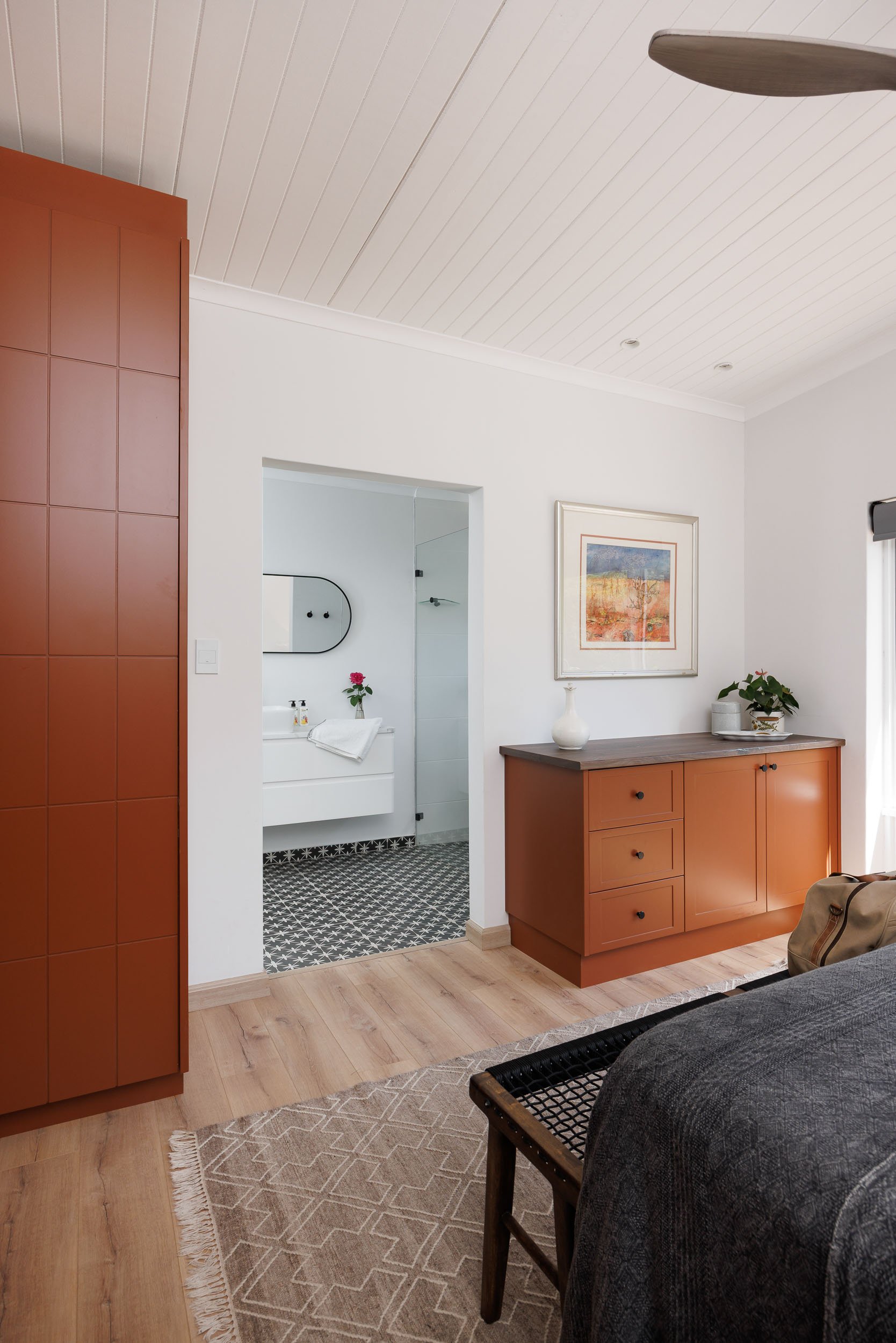 A cozy room with a wooden sideboard, a framed artwork on the white wall, and a table with a vase and plant. An open doorway reveals a bathroom with black and white patterned floor tiles, a sink with a toothbrush and a single rose in a vase. Part of a