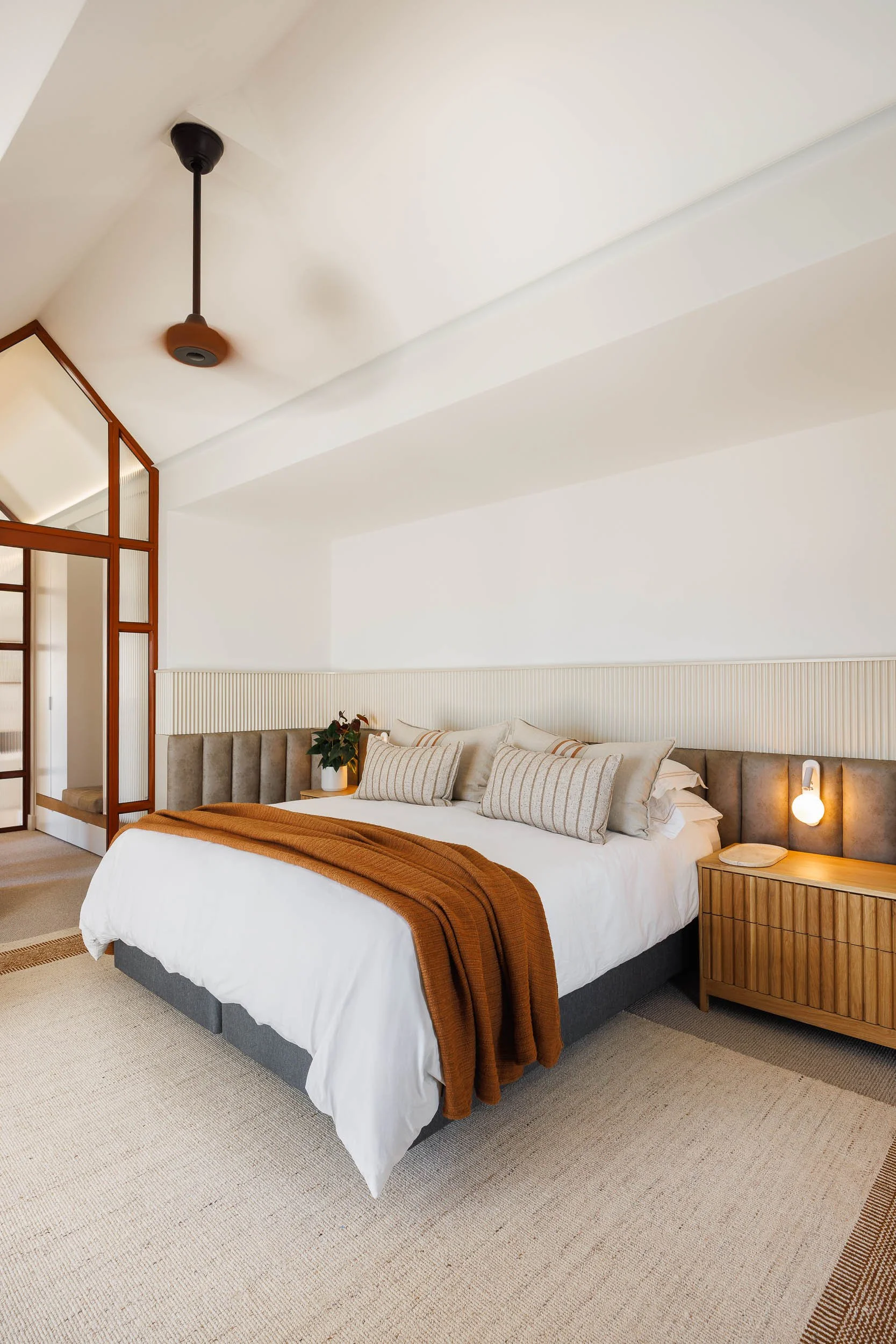 Master bedroom with pitched ceiling, channelled velvet headboard, amber throw, and timber bedside drawers