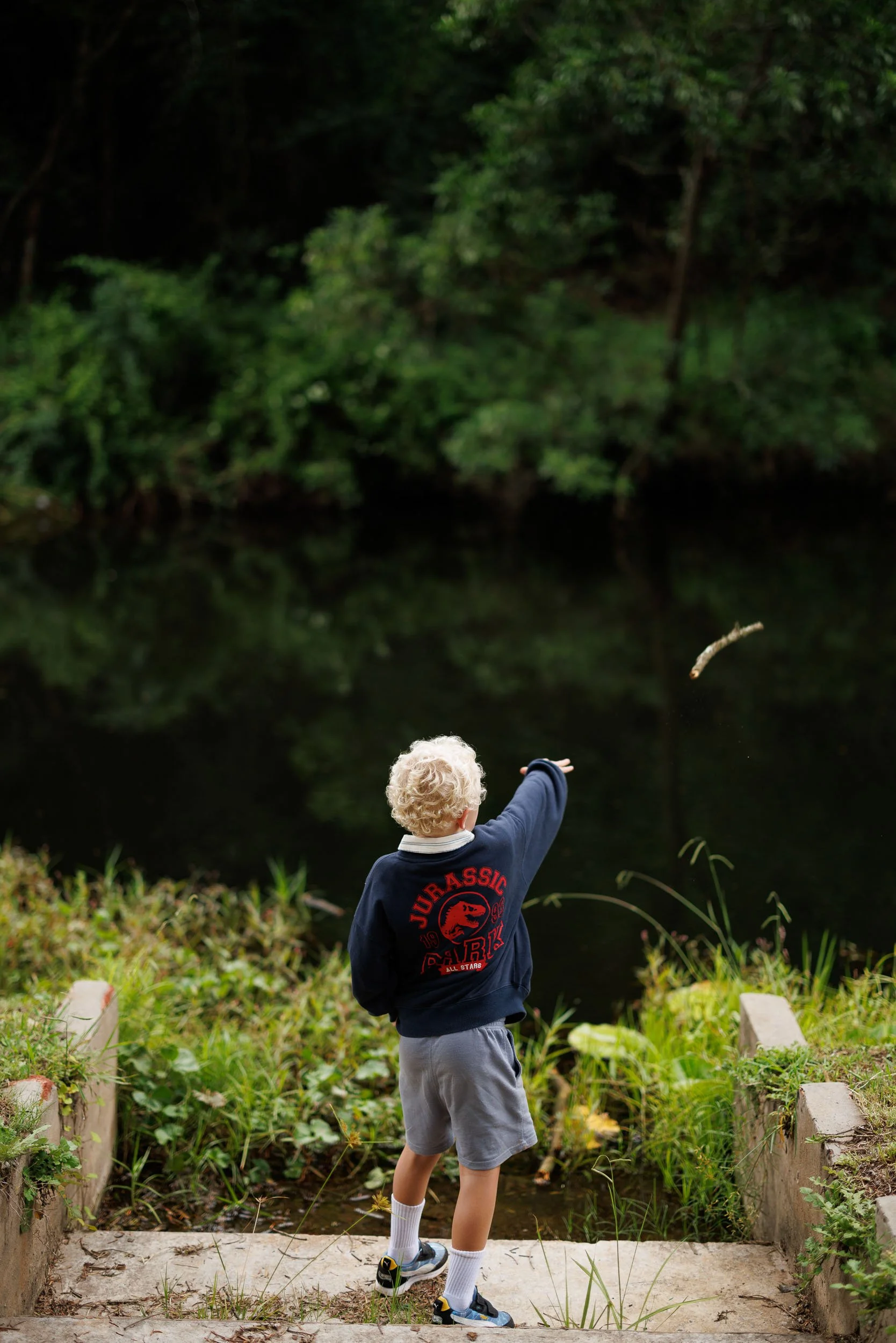 A young child with curly blond hair, wearing a navy blue hoodie with a Jurassic Park logo, gray shorts, white socks, and sneakers, standing on a concrete step by a small creek, throwing a stick into the water. The background features green trees and 