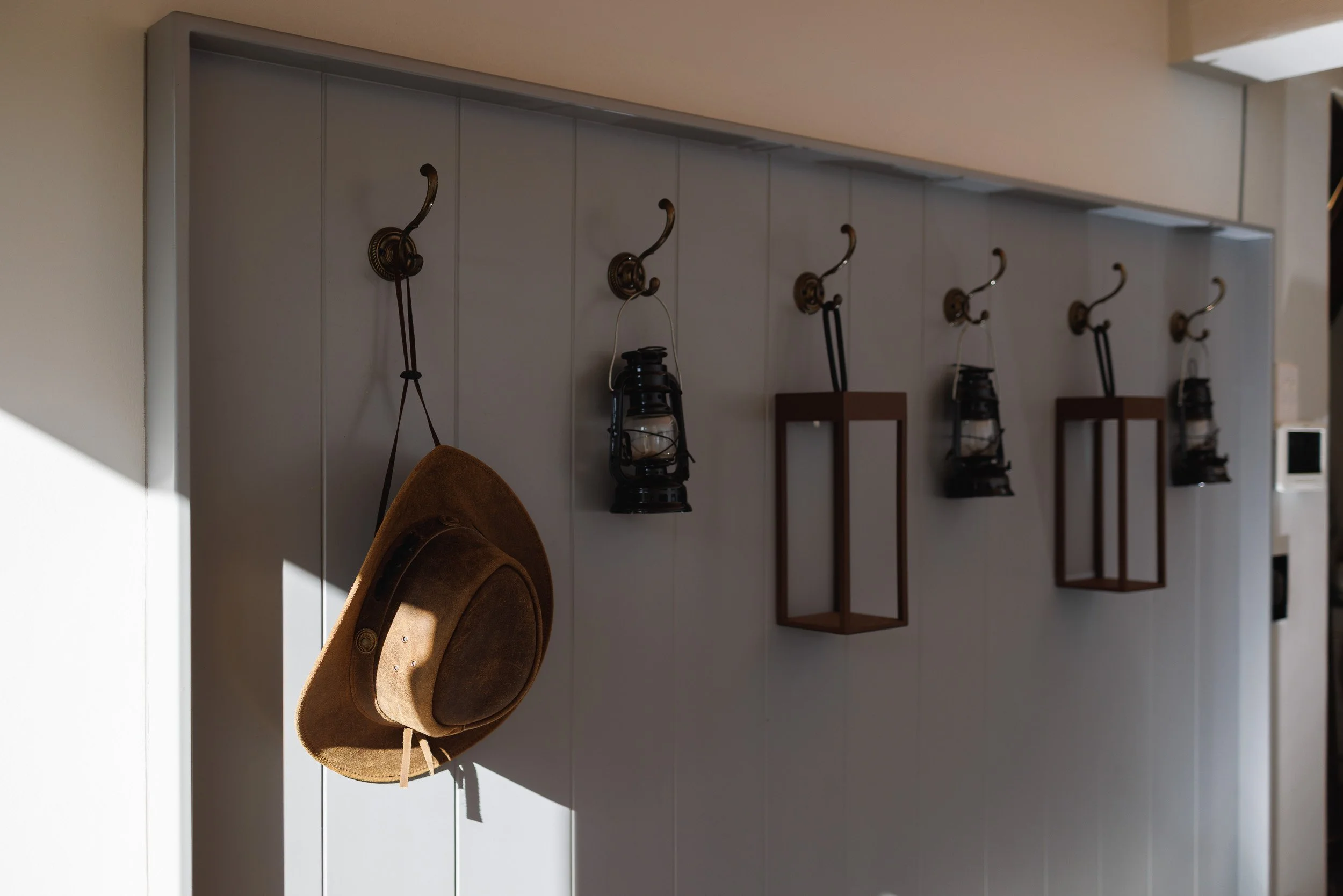 A wall with four brass hooks, two black lanterns hanging from two hooks, a brown frame, and a brown hat hanging on a hook. The wall is gray, and there's a small white device on the right side.