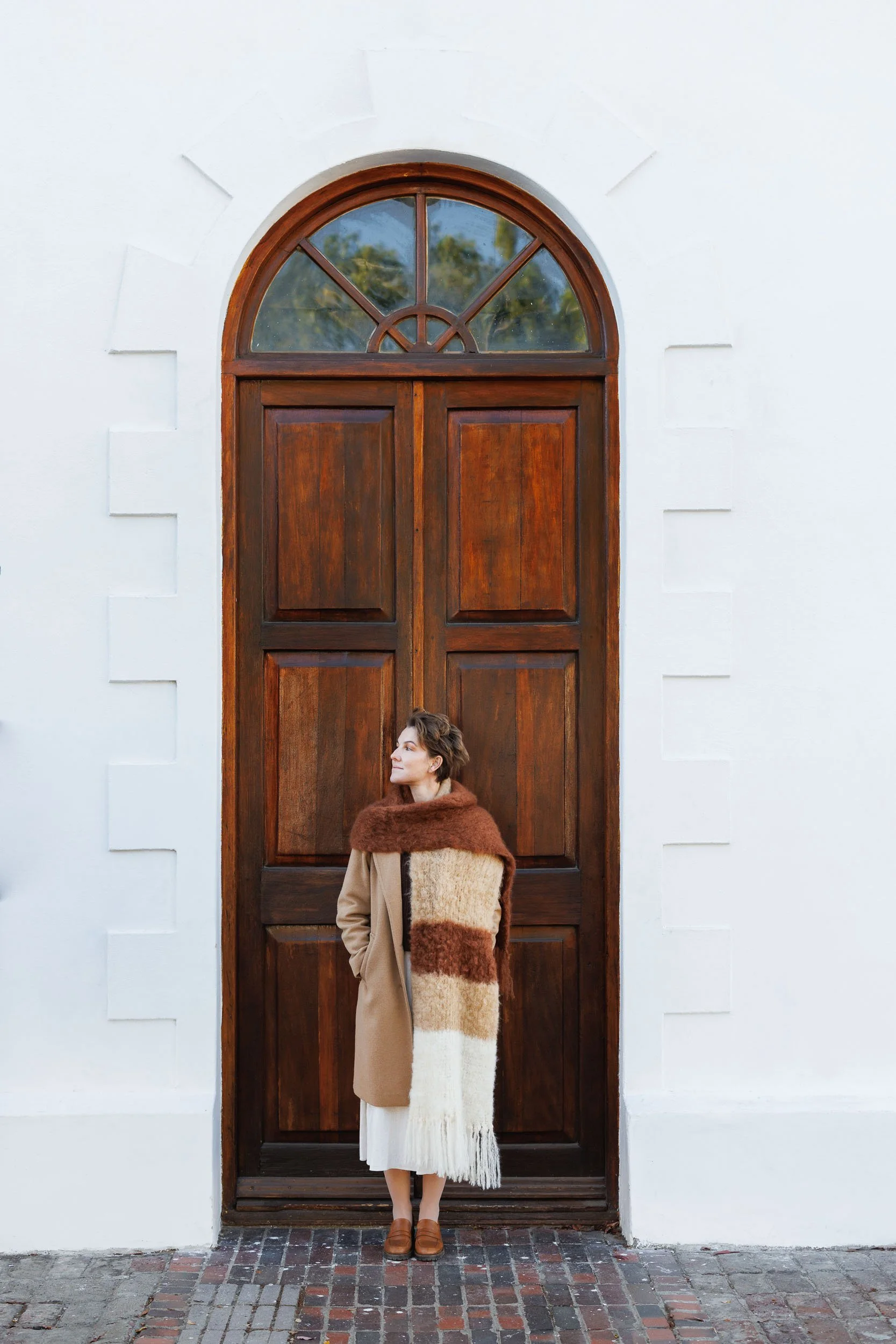 A woman in a beige coat with a large, fuzzy, color-blocked scarf stands in front of a wooden door with an arched window. The door is set in a white wall with decorative brickwork at the base and around the edges. The woman is looking to her left.