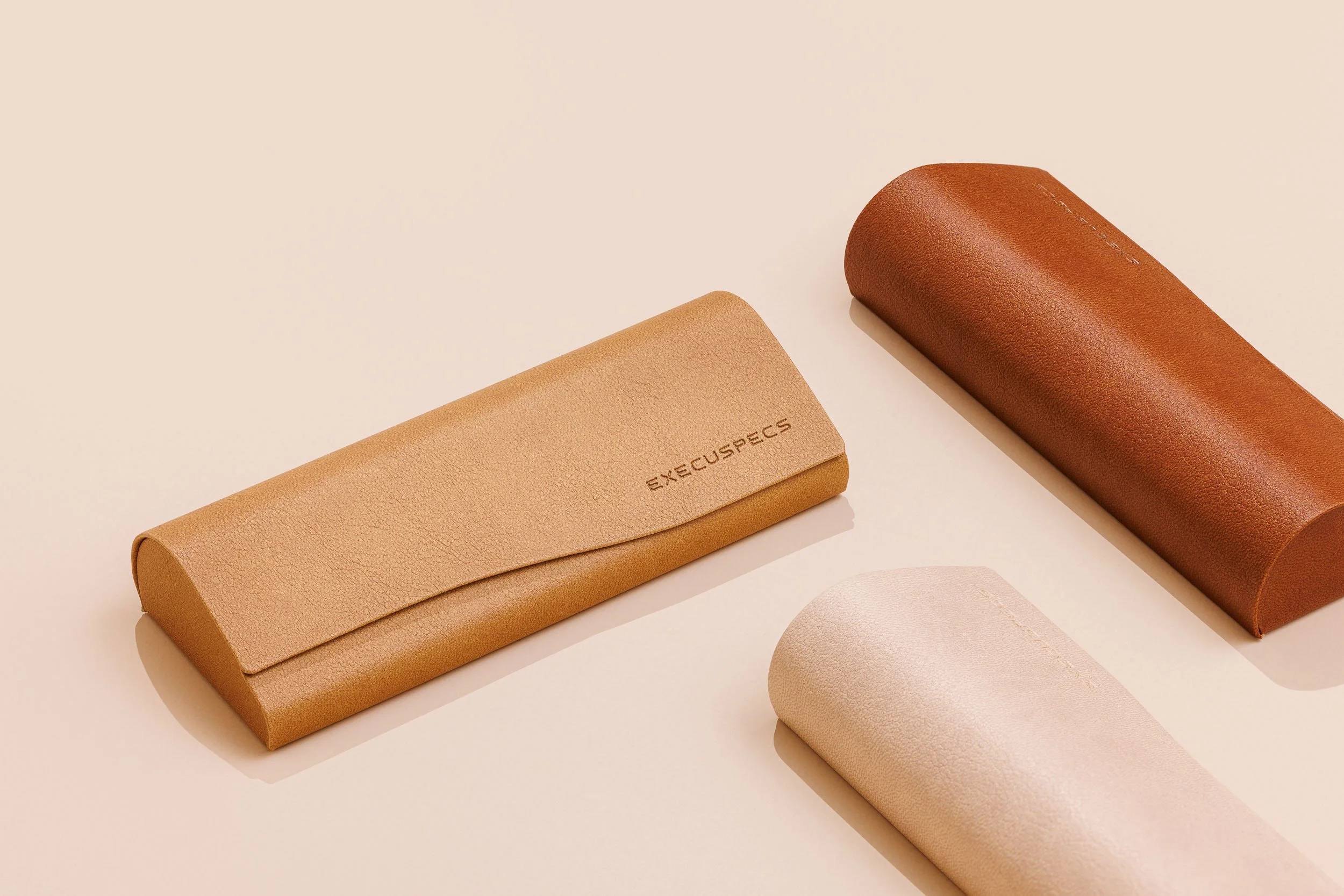 Two brown leather eyeglass cases and a pinkish-beige round case on a light beige background