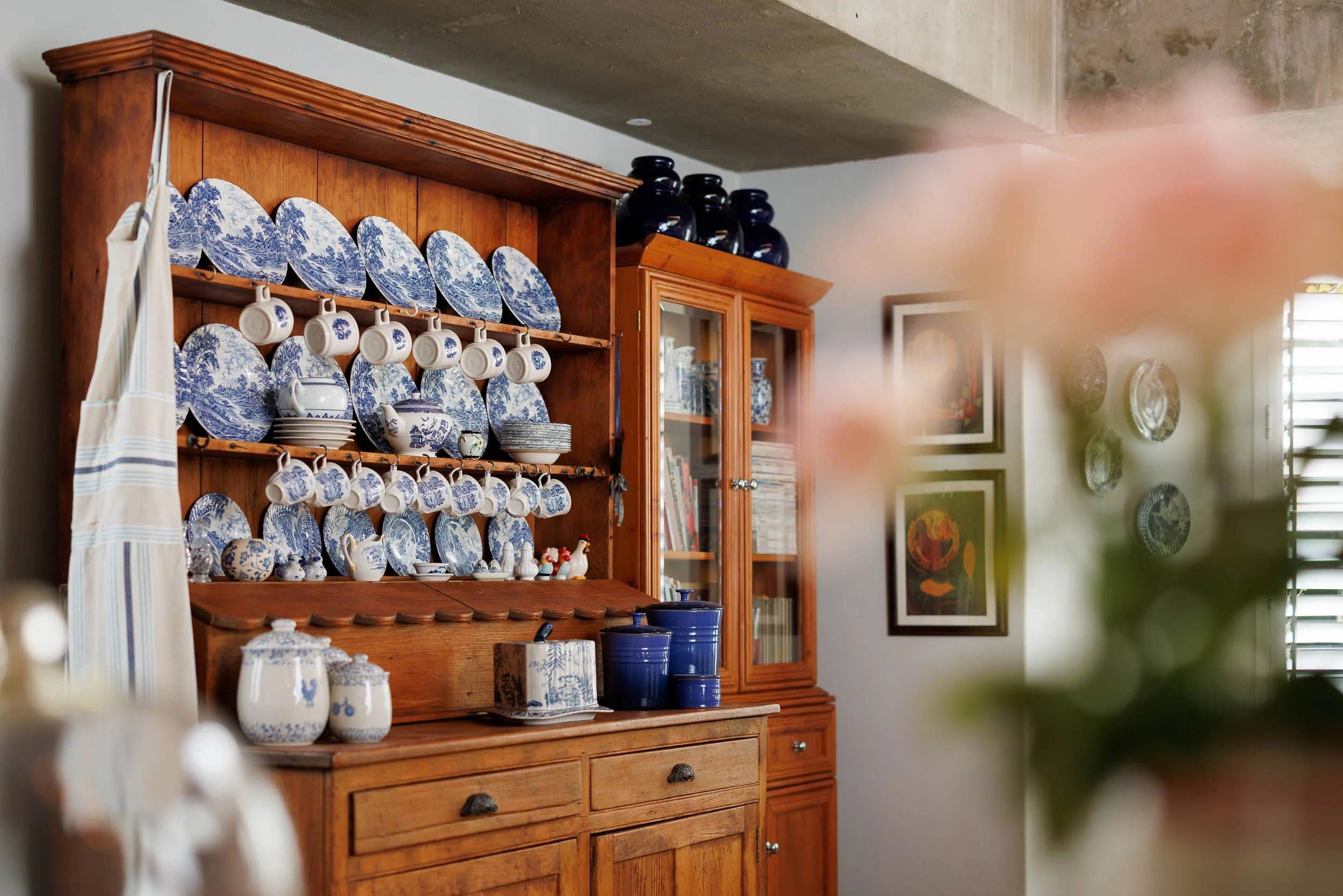A wooden china cabinet displaying blue and white decorative plates, cups, and bowls, with a glass-fronted section containing books and other items. Multiple black vases are on top of the cabinet, and framed pictures are on the wall to the right.
