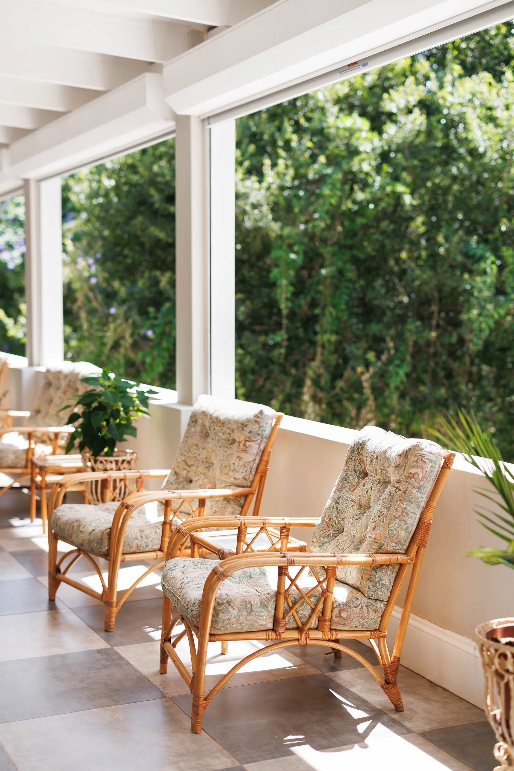 Sunlit porch with wicker chairs with floral cushions, green foliage outside, and tiled floor.