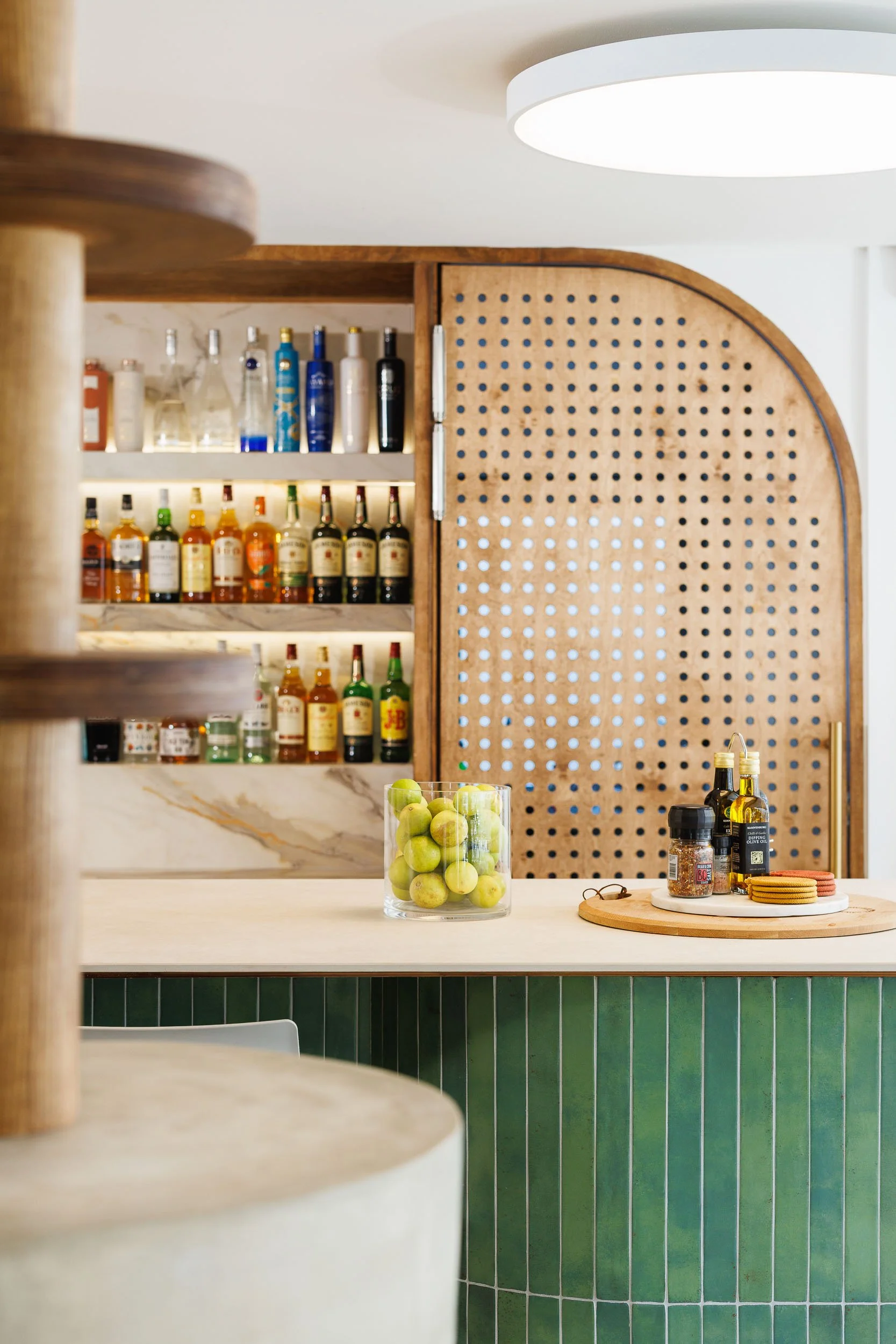 Bar nook with green subway tile island, arched perforated timber cabinet door, and backlit liquor shelves