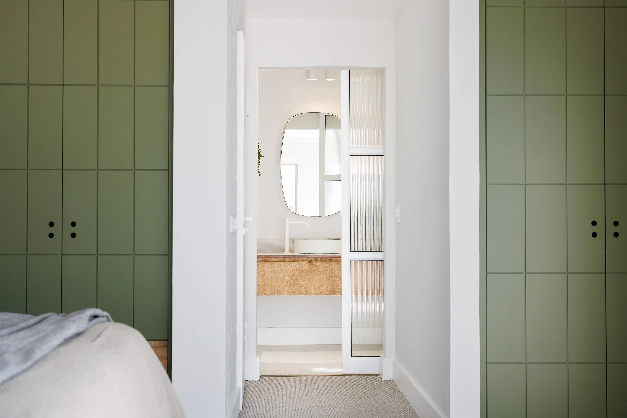 Sage green grid-panel wardrobes flanking hallway leading to ensuite with oval mirror and reeded glass door