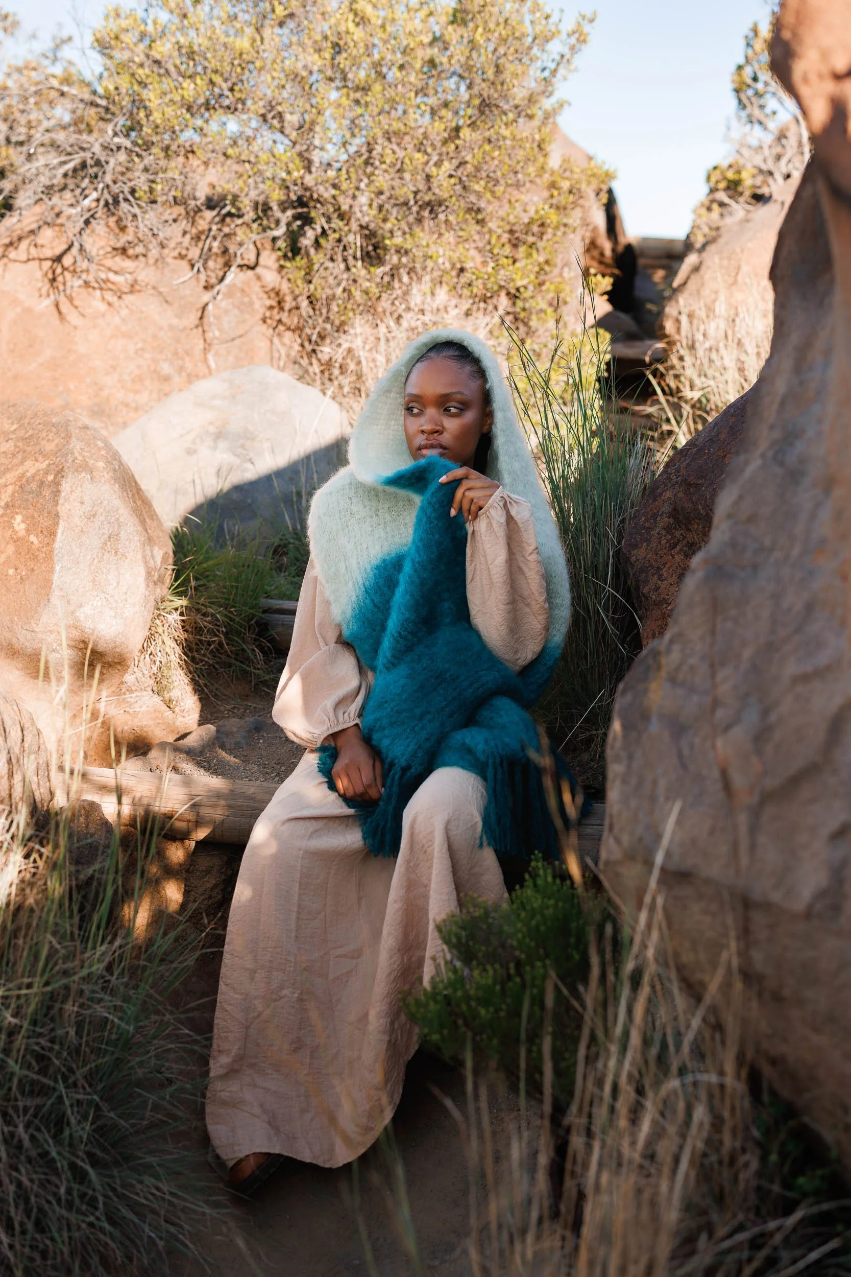 A woman sitting outdoors among rocks and plants, wearing a beige dress, a teal and cream shawl, and a hood; she has dark skin and is looking to the right with a contemplative expression.