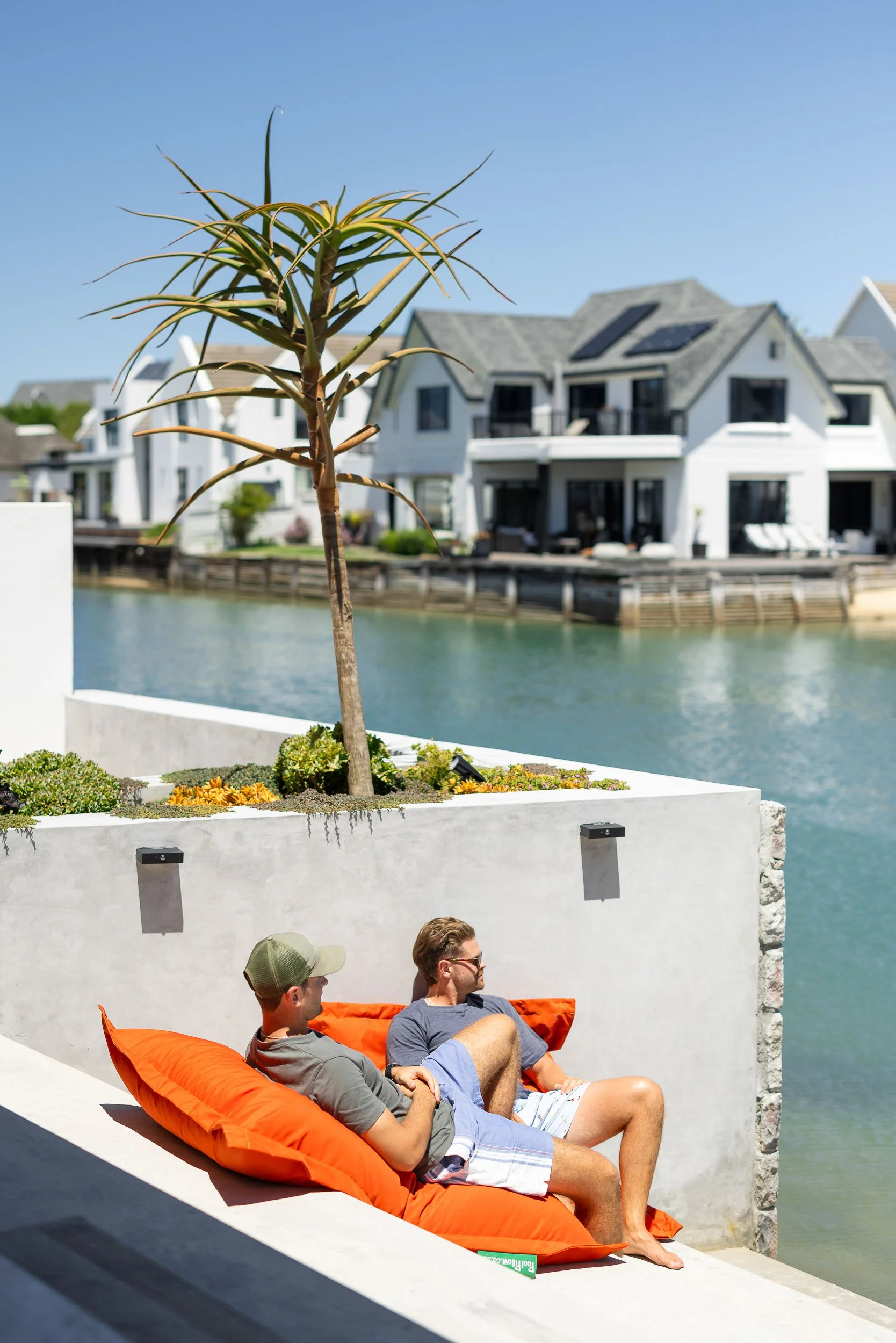 Two people relaxing on orange bean bags on a canal-side terrace with water and neighbouring homes in background