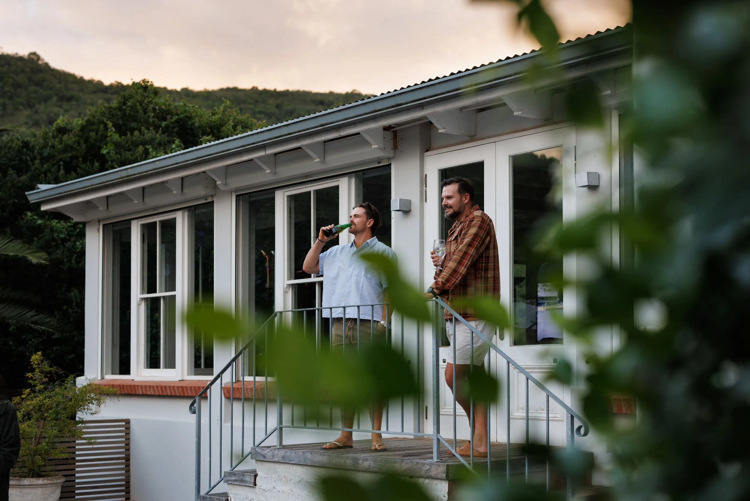 Two men standing on a small balcony outside a white house, drinking beverages and smiling, during sunset. Green bushes and distant hills in the background.