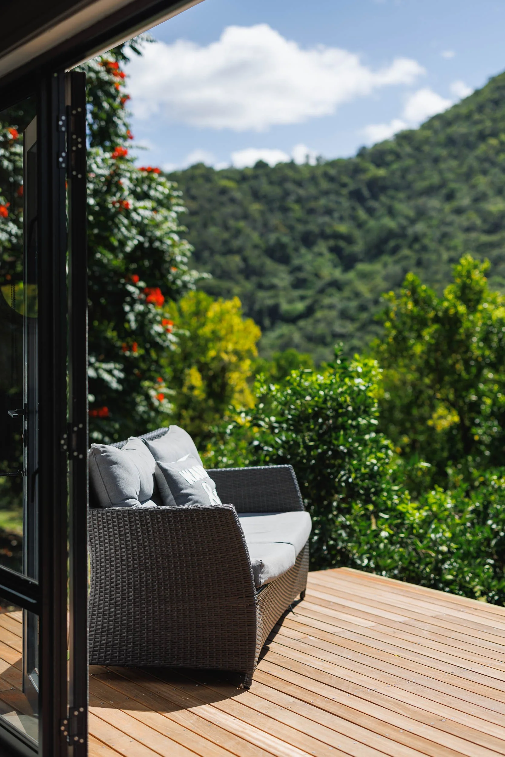 Outdoor terrace with wicker couch and cushions overlooking lush green hillside and cloudy sky.