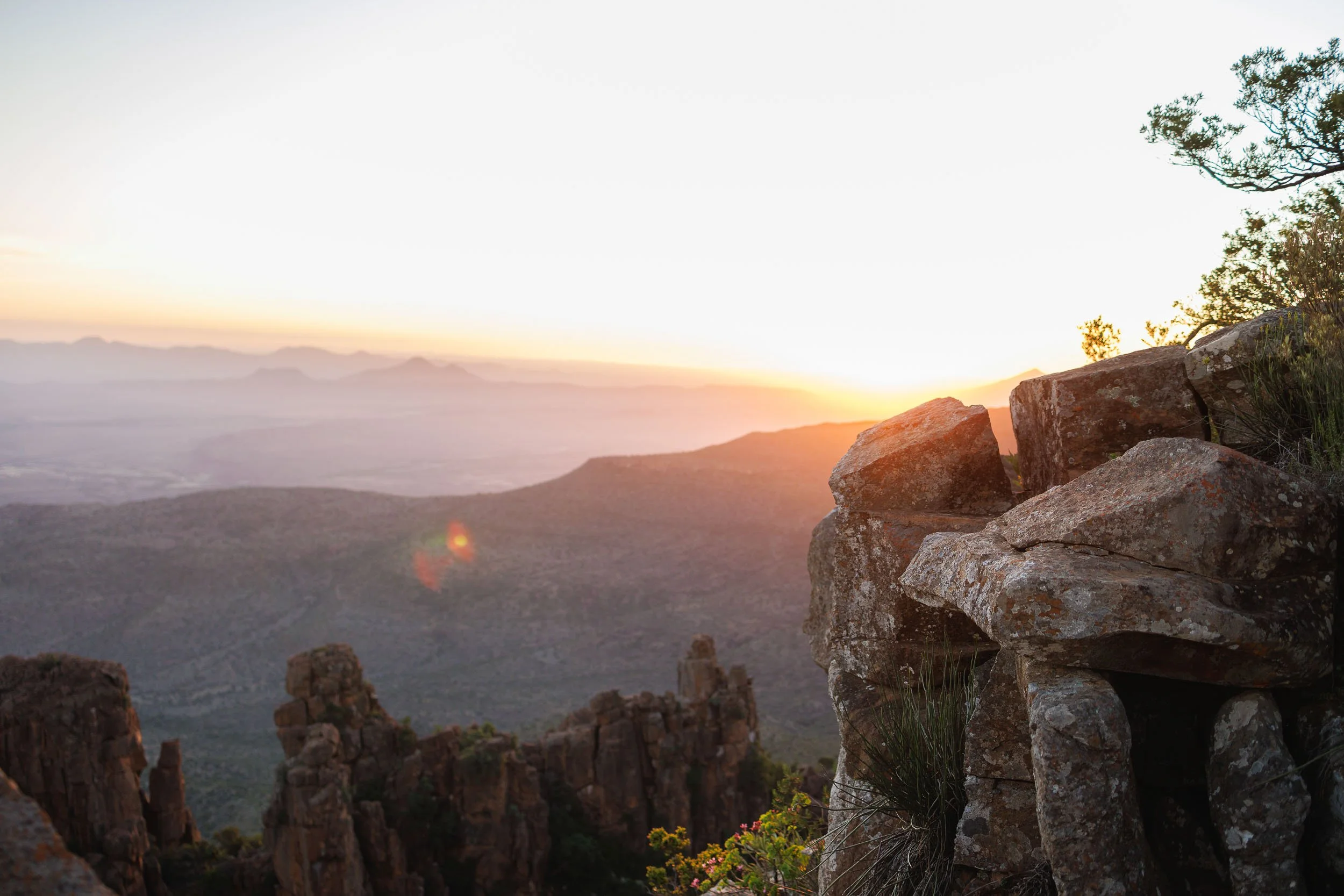 A scenic view of a mountain landscape at sunrise, with rugged rocks and sparse vegetation in the foreground, and distant mountain ranges under a bright sky.