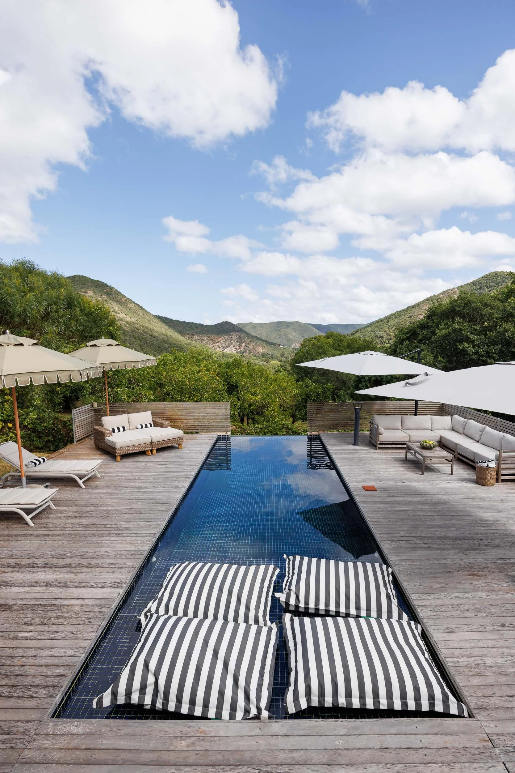 A swimming pool with two striped cushions floating in it, surrounded by a wooden deck with outdoor furniture and umbrellas, set against green hills and a blue sky with scattered white clouds.