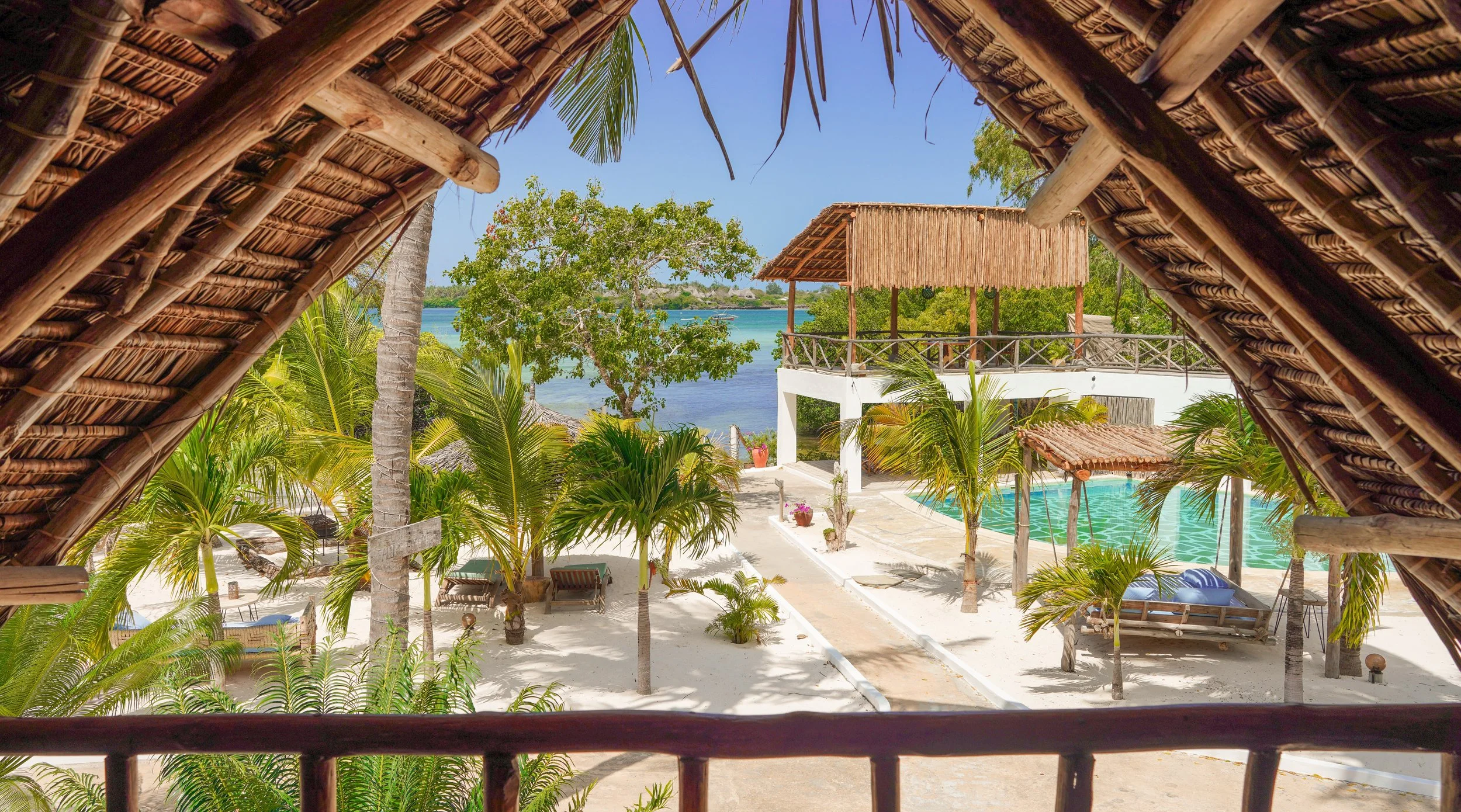 View from a thatched roof hut overlooking a tropical resort with palm trees, a pool, and a view of the water and sky.