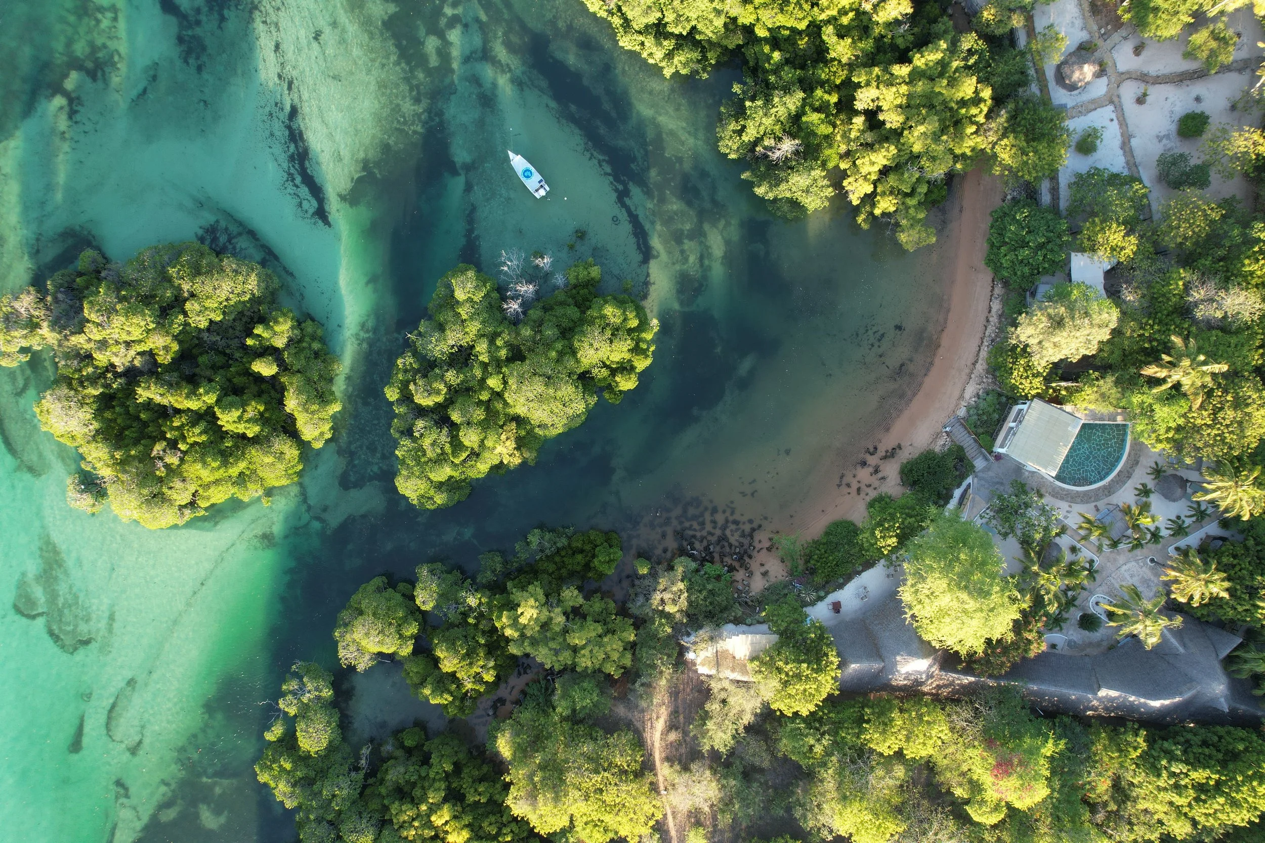An aerial view of a coastal area featuring clear turquoise waters, lush green trees, a sandy beach, and some buildings and structures near the shore.