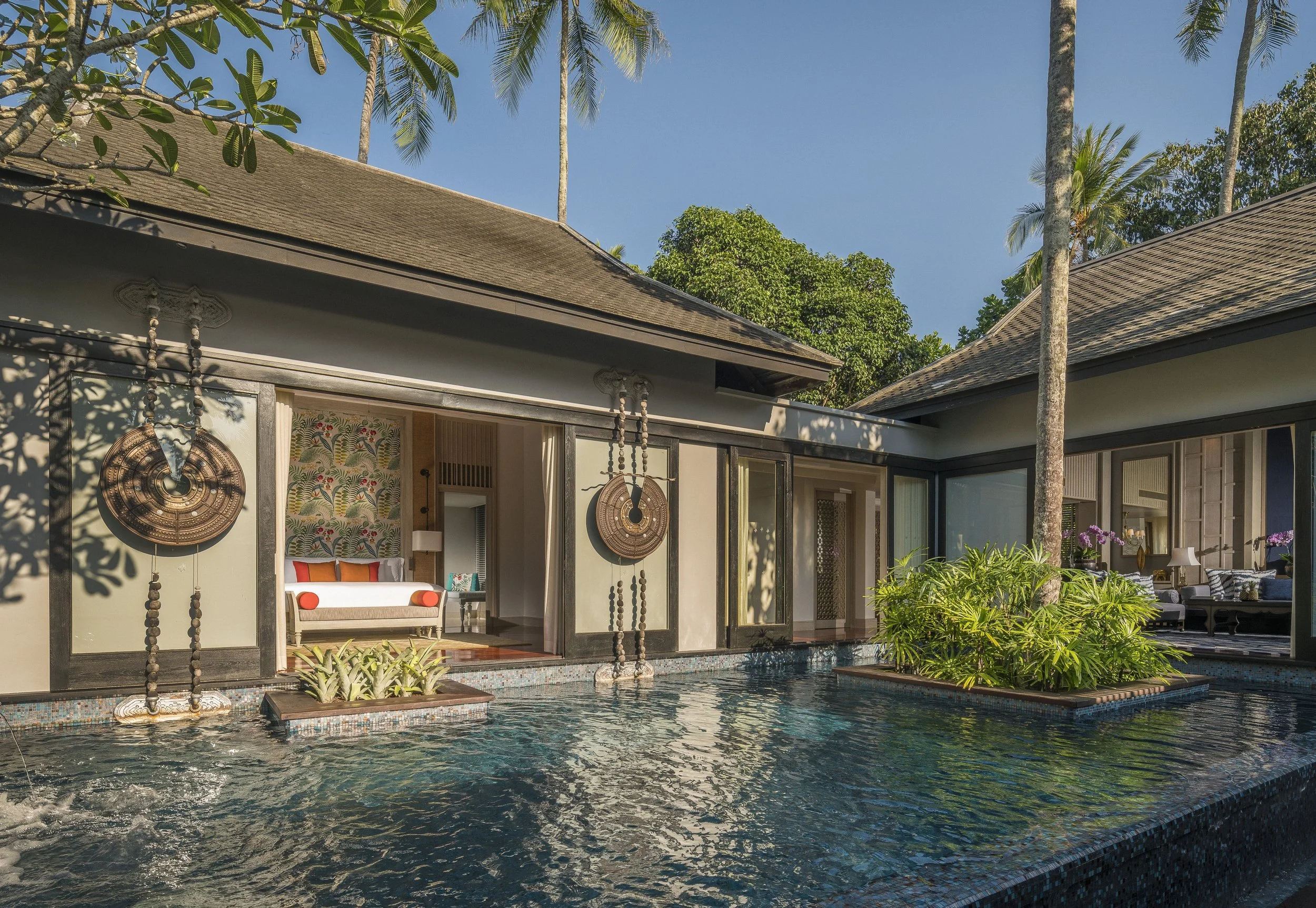 A view of a house with an open living area overlooking a pool with aquatic plants, surrounded by trees and greenery, under a clear blue sky.