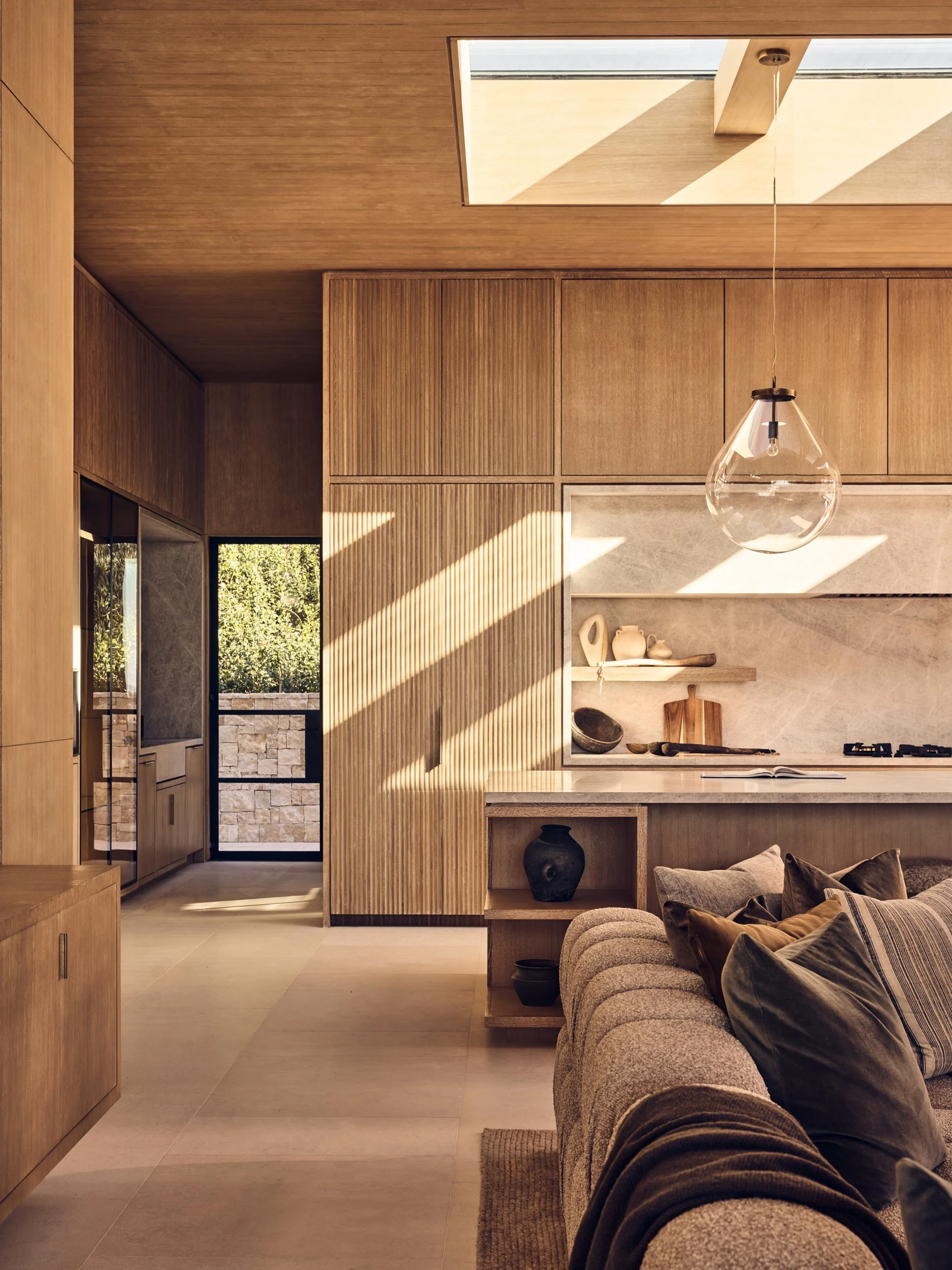 A modern interior with wooden cabinetry, a kitchen area with open shelves, and a couch with pillows. Natural light streams through a skylight and window, casting shadows on the wall.