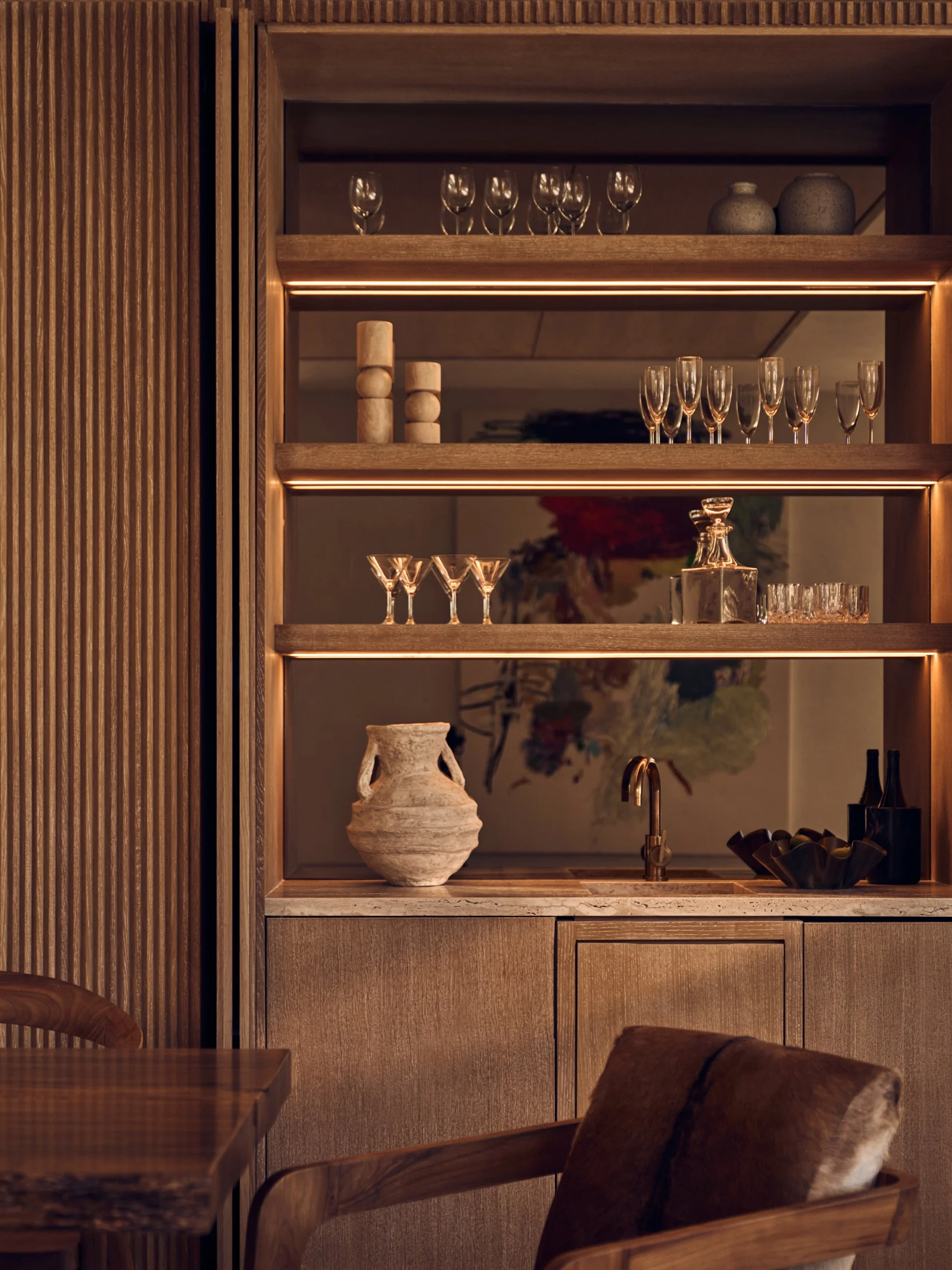 A wooden shelving unit with glassware and decorative items, illuminated by warm lighting, with a stone vase and a painting of flowers in the background.